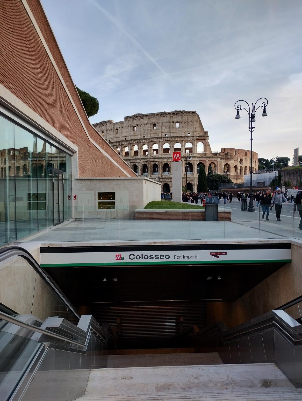 In primo piano l'entrata della stazione della metropolitana "Colosseo fori imperiali", con scala al centro e scale mobili ai due lati, l'insegna più un là e sullo sfondo il Colosseo