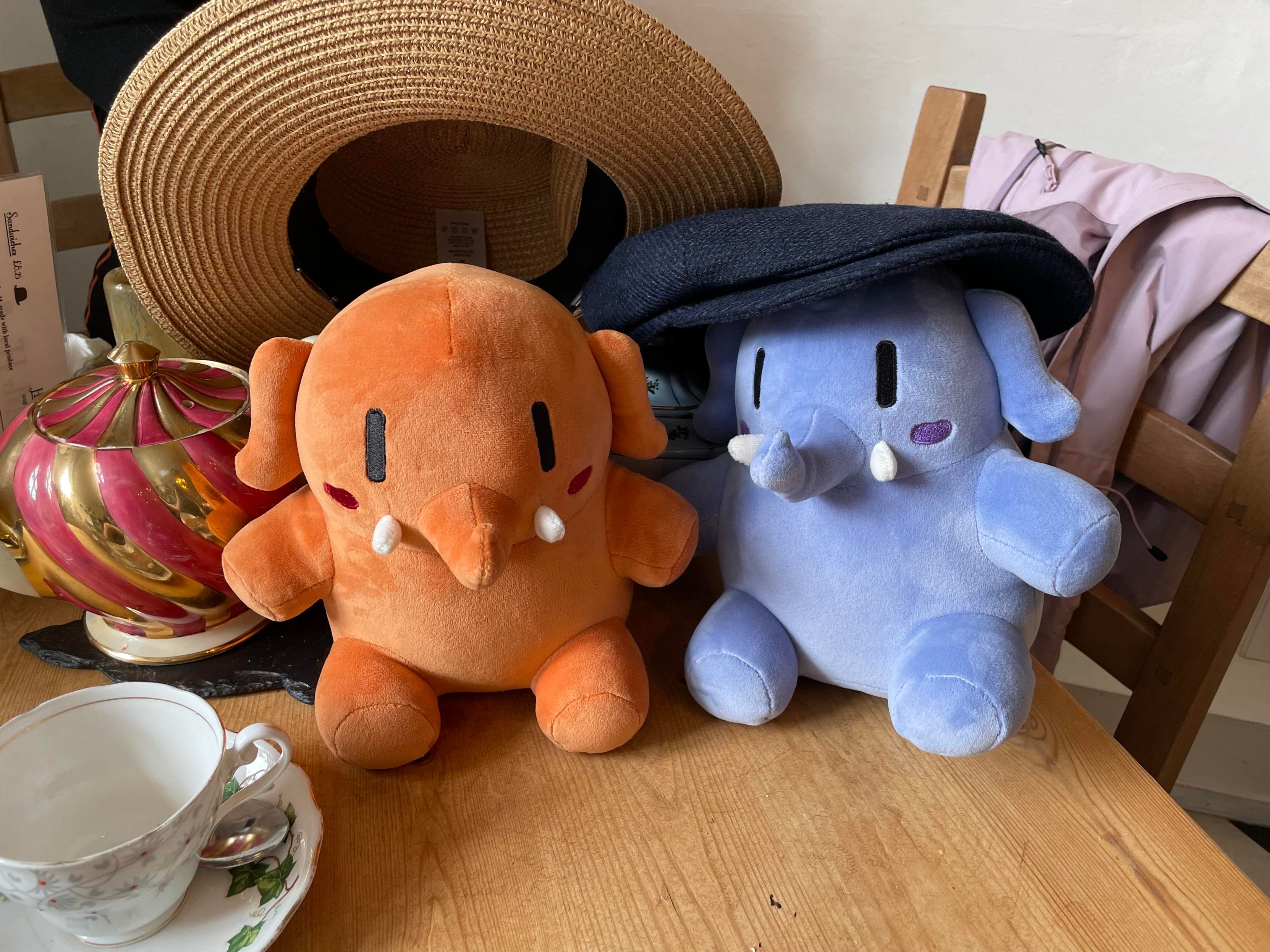 An apricot colored and a blue Mastodon stuffed toys sitting on a table next to each other. The blue one is wearing a human sized blue hat, and the apricot one a straw hat. 
