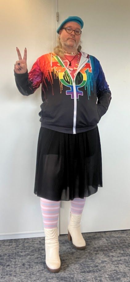 Full body selfie of myself. I'm wearing a black hoodie with a rainbow drip motif across the shoulders and sleeves and a large rainbow transgender symbol in the middle. It is partly zipped open, revealing a pink t-shirt underneath, which pairs with white blockheel boots and a light blue beret to form the colours of the transgender flag. Under my over-the-knee black summer skirt, I'm wearing tights with bands in the colours of the transgender pride flag.