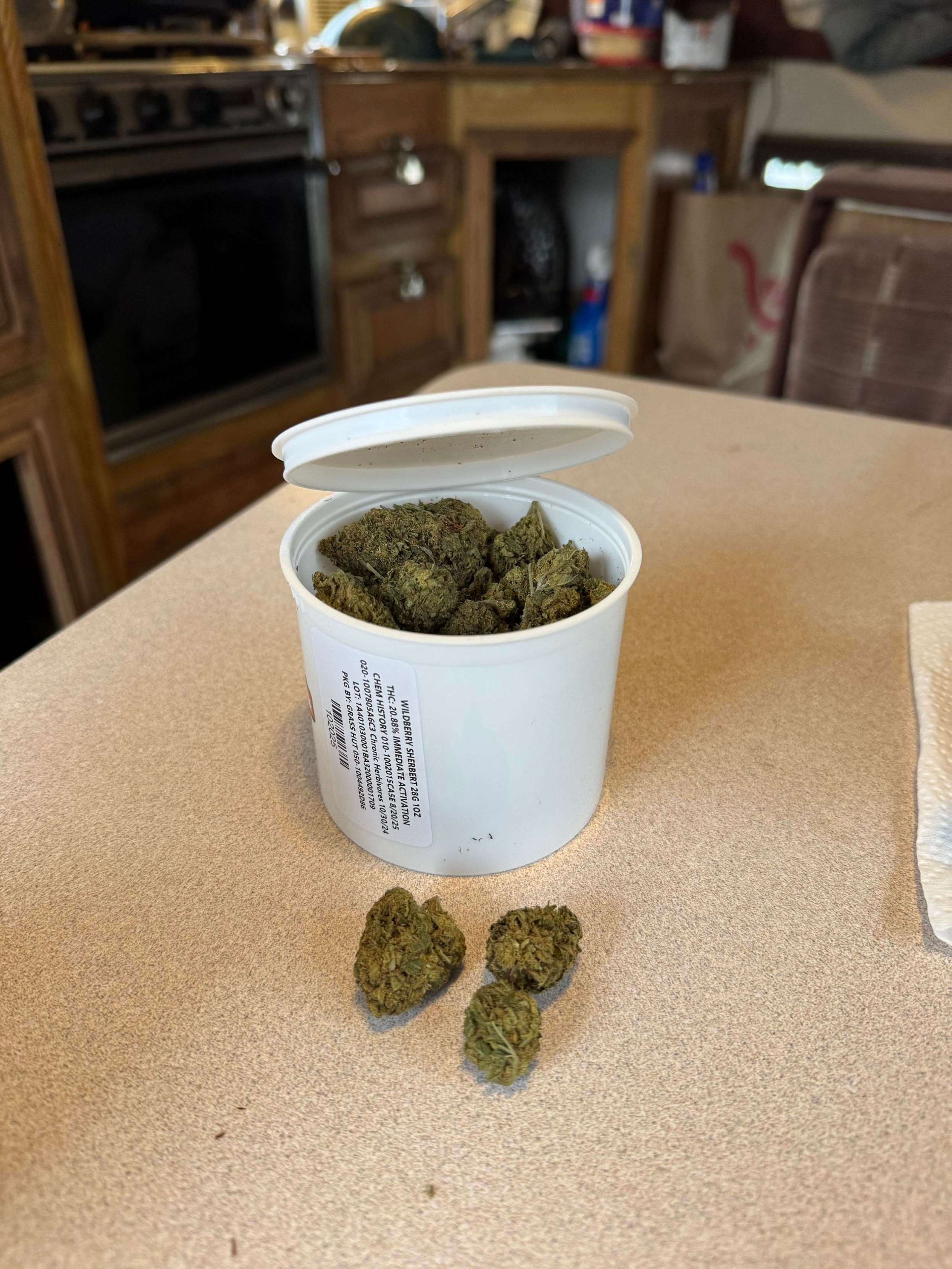 A white container is open, revealing cannabis buds inside. Three additional buds are placed in front of the container on a table. In the background, there is a kitchen with a stove and cabinetry.