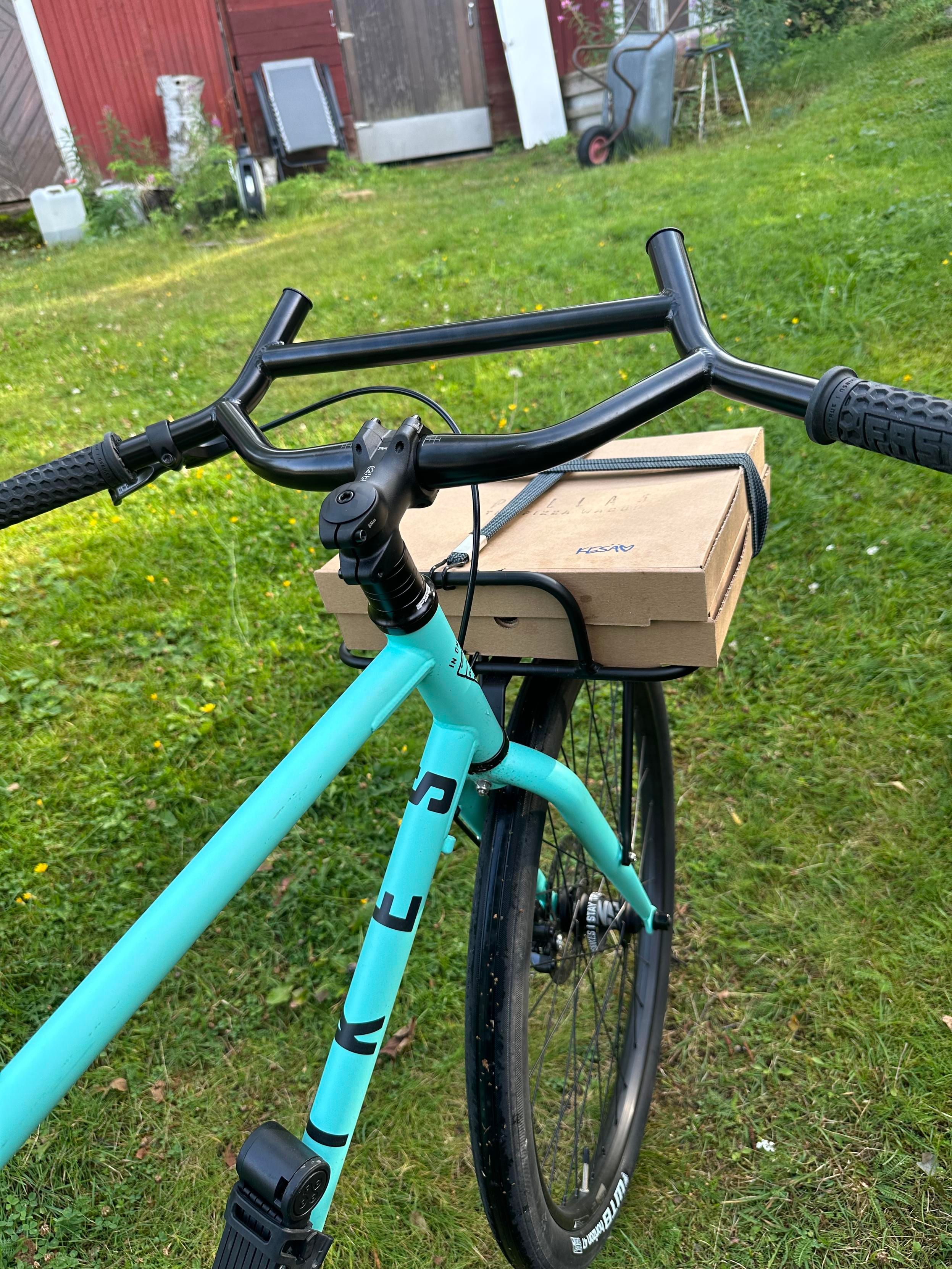 Sama mintunvihreä fillari vihreää nurmikkoa vasten. Etutarakalla kaksi pizzalaatikkoa littanoilla mustekaloilla kiinnitettyinä. Taustalla punainen piharakennus, ja sekalaista krääsää kuten kottikärryt, pihatuoli ja jakkara