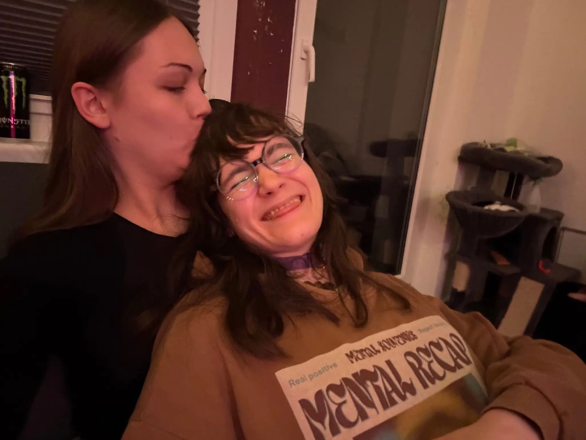 Photo of two adorable feminine individuals in an affectionate moment. The person on the right is leaned onto the woman on the left, while the woman on the left is giving the person a kiss on their forehead. The person on the right is visibly very giddy about this and is making a cute, very happy face, with their eyes closed and sticking their tongue out cheekily. Both are sitting on a couch in warm, moody lighting