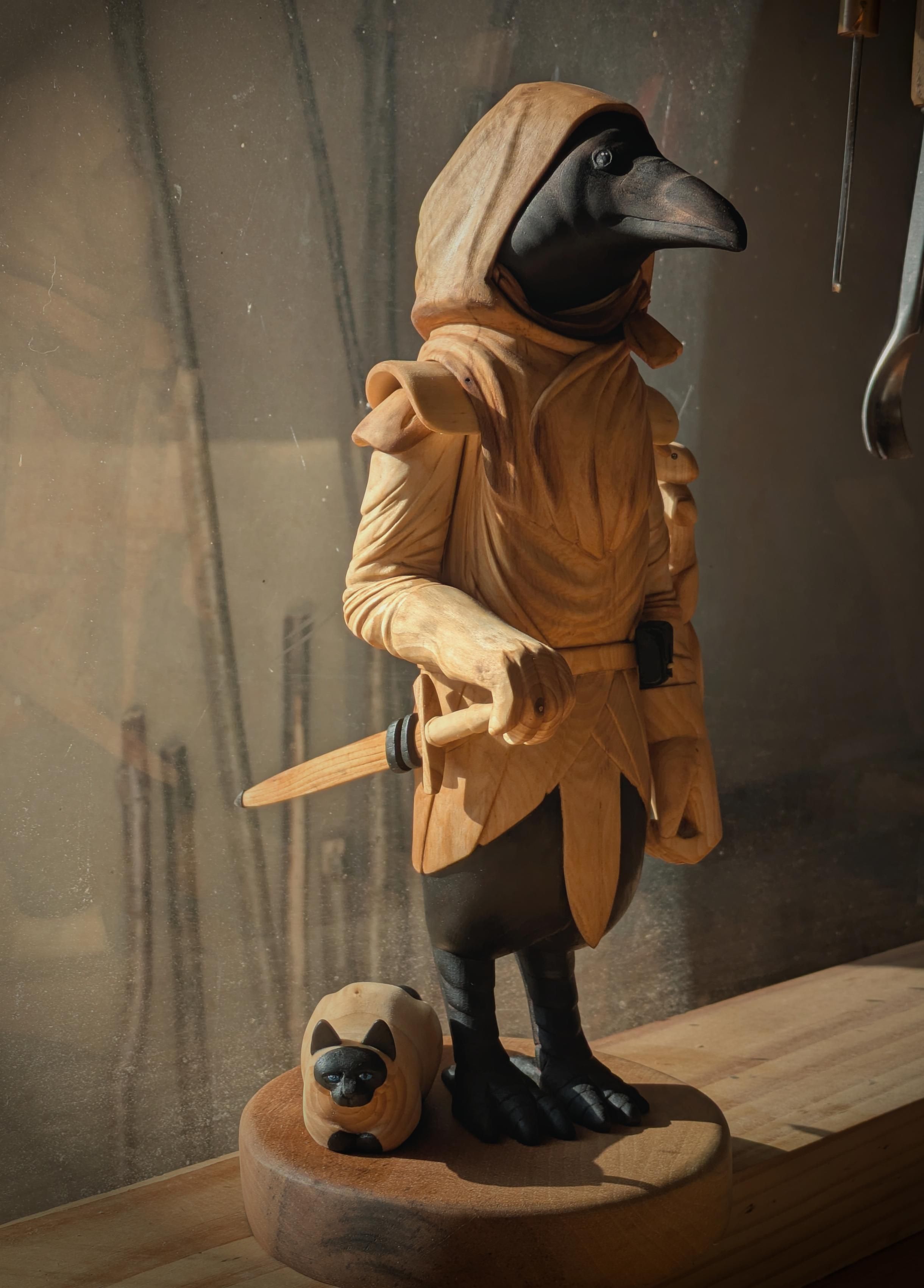 Woodcarved humanoid crow with a hooded cloak, a sword, satchel on its belt. It has a squirrel climbing its arm, and a cat sitting next to its feet