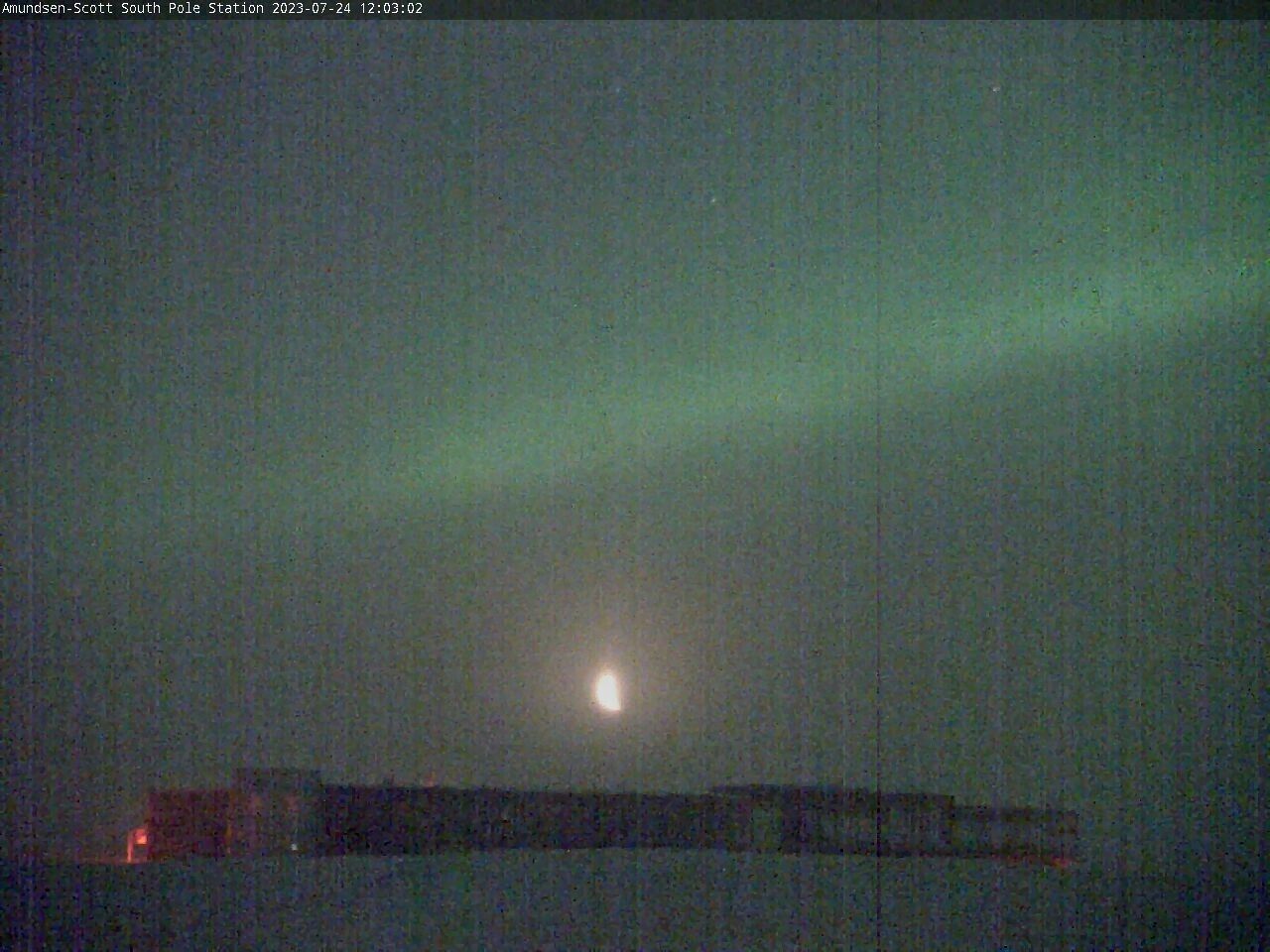 A grainy night-time webcam picture of a research station at the South Pole, a large building low to the ground.

Above it is a bright green light, across the night sky, and right between it and the research building is a brightly lit up crescent moon.