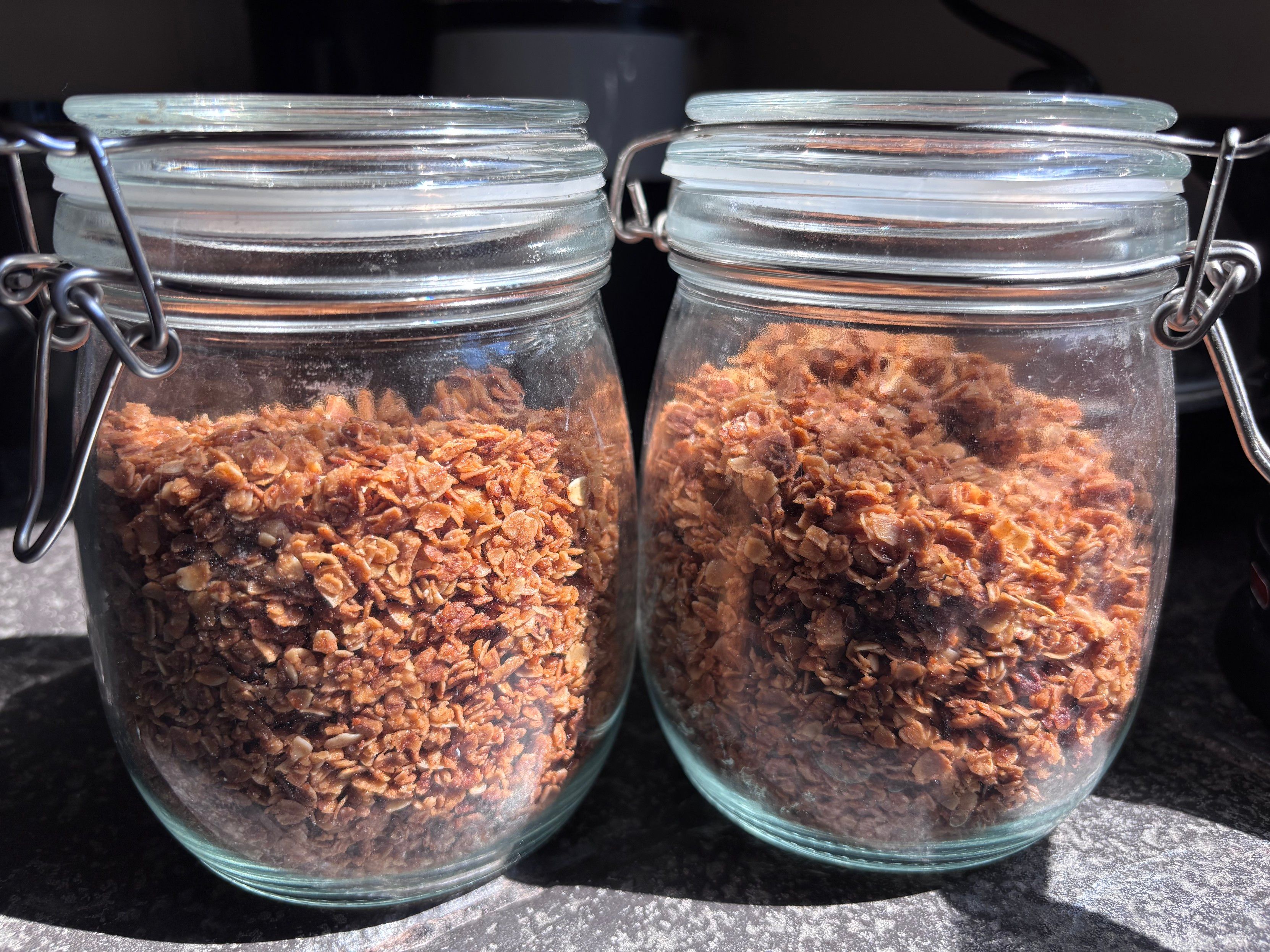 Two glass jars filled with granola, placed on a countertop with sunlight illuminating them.