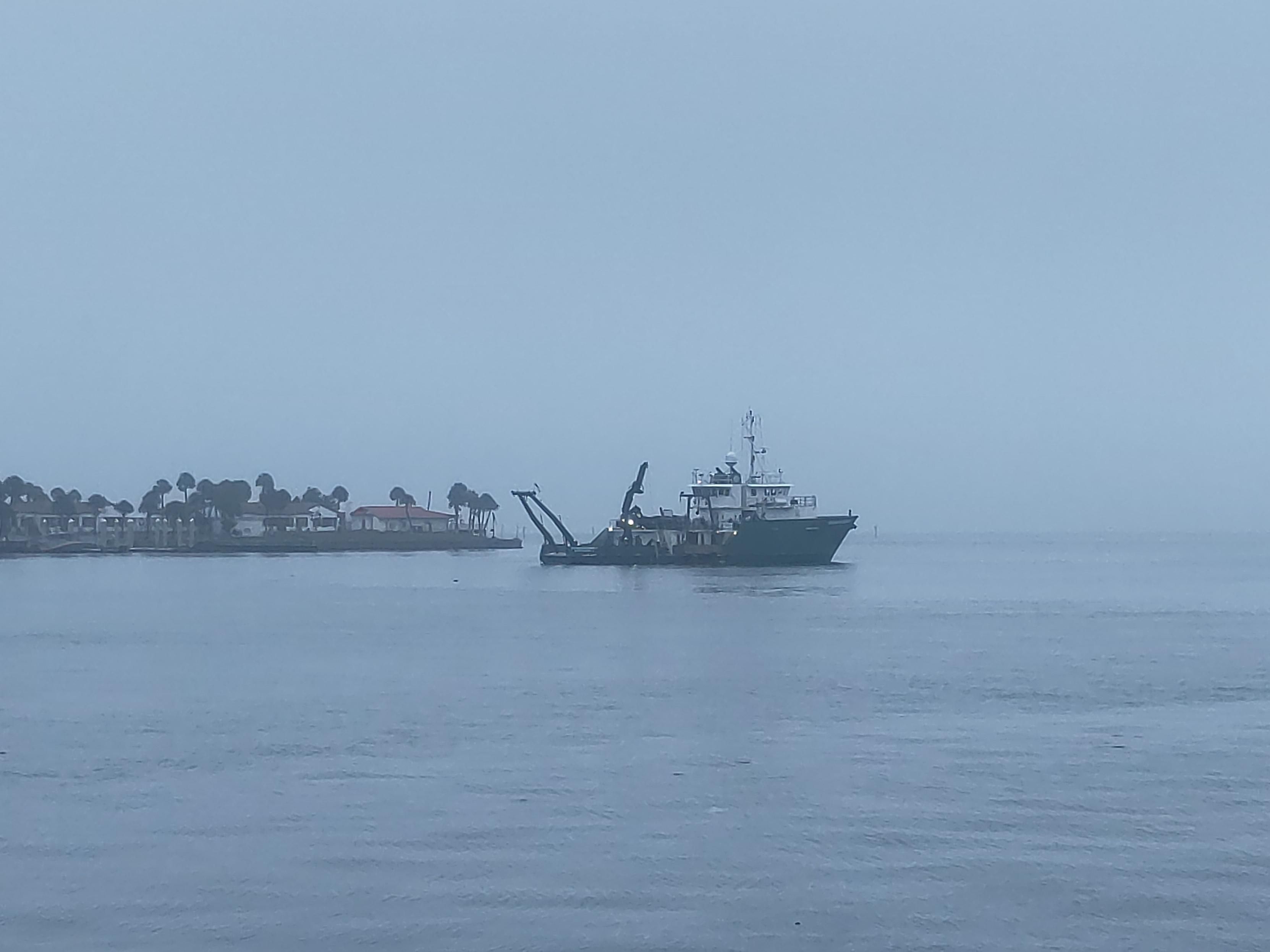 A green-hulled research vessel, the RV Weatherbird, with its bow toward the right and its stern toward the left. The A-frame, a large moveable frame attached to the stern, hangs out sub-vertically from the stern, over the flat calm, gray water. Lights shine in various places on the ship. The quarters and bridge are white, in contrast to the dark green hull which looks black in the photo. There is hardly any visible horizon due to overcast foggy conditions. There is a small peninsula to the left (aft) of the ship, with white buildings that have read tile grooves and several palm trees which are taller than the buildings. 