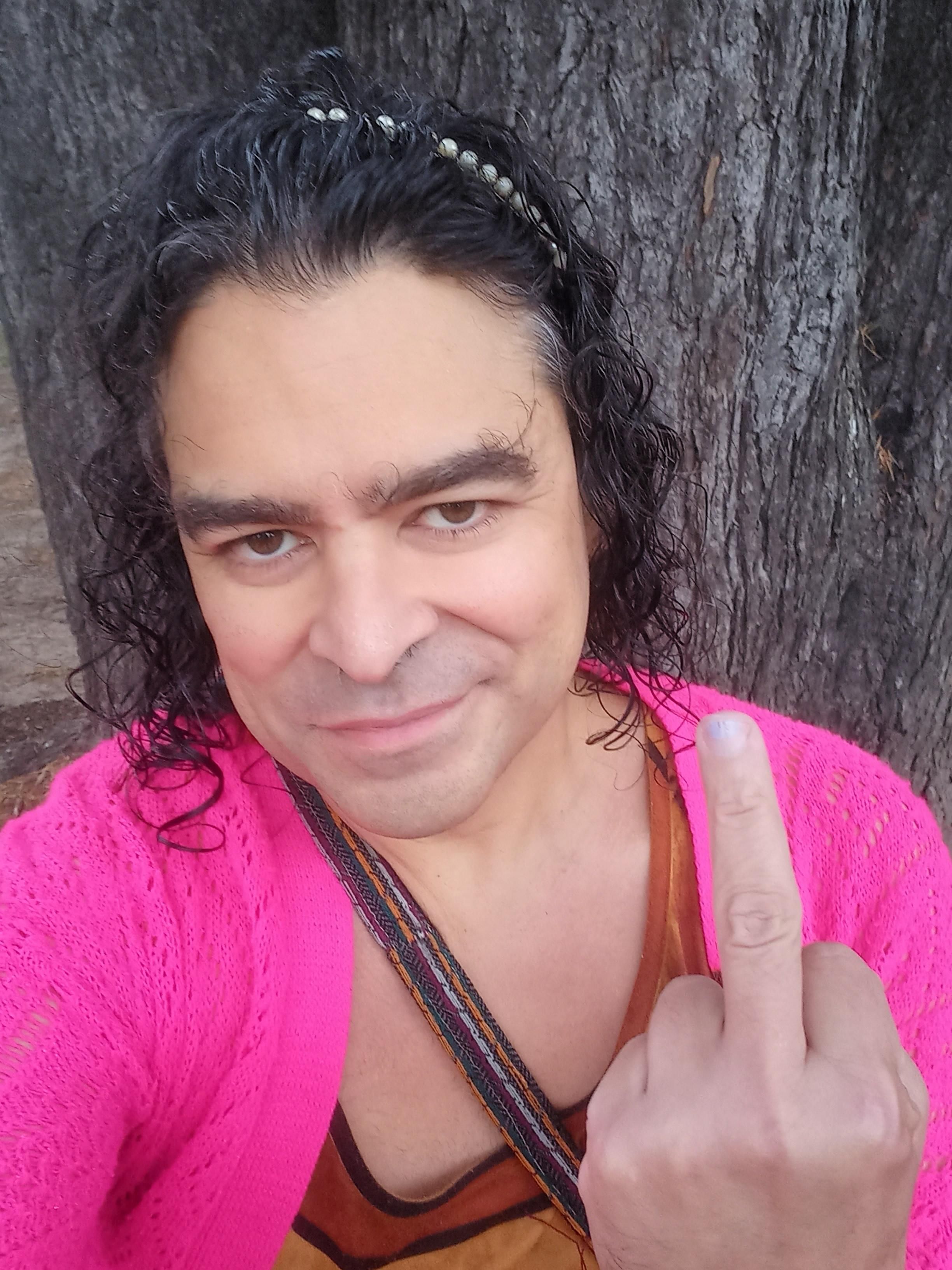 Yours truly grinning at the camera and giving the finger. Wet black hair held back by a wire band with cheap plastic pearls, dress in earth tones, pink open weave cardigan, strap across my chest. Grey bark of a big, old tree behind me.