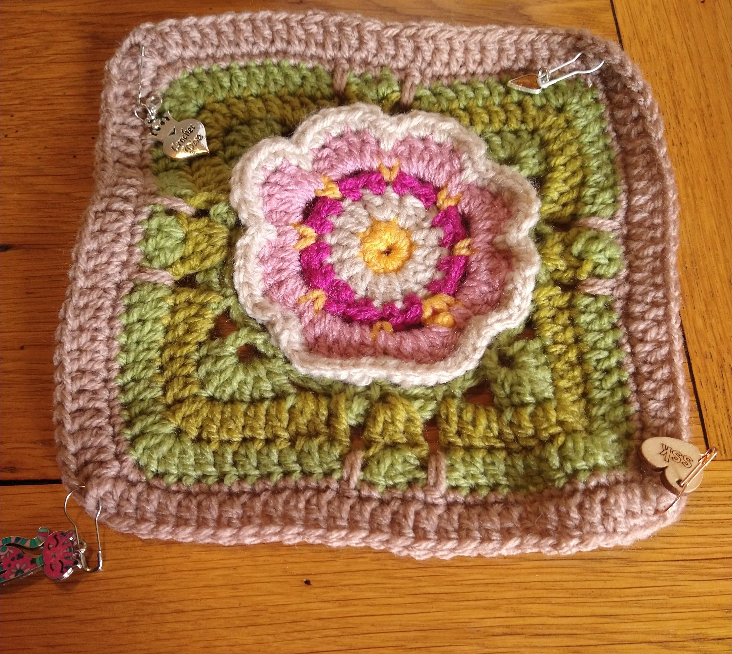 One crochet square with a pink lilly like flower in the centre of a green and beige background 