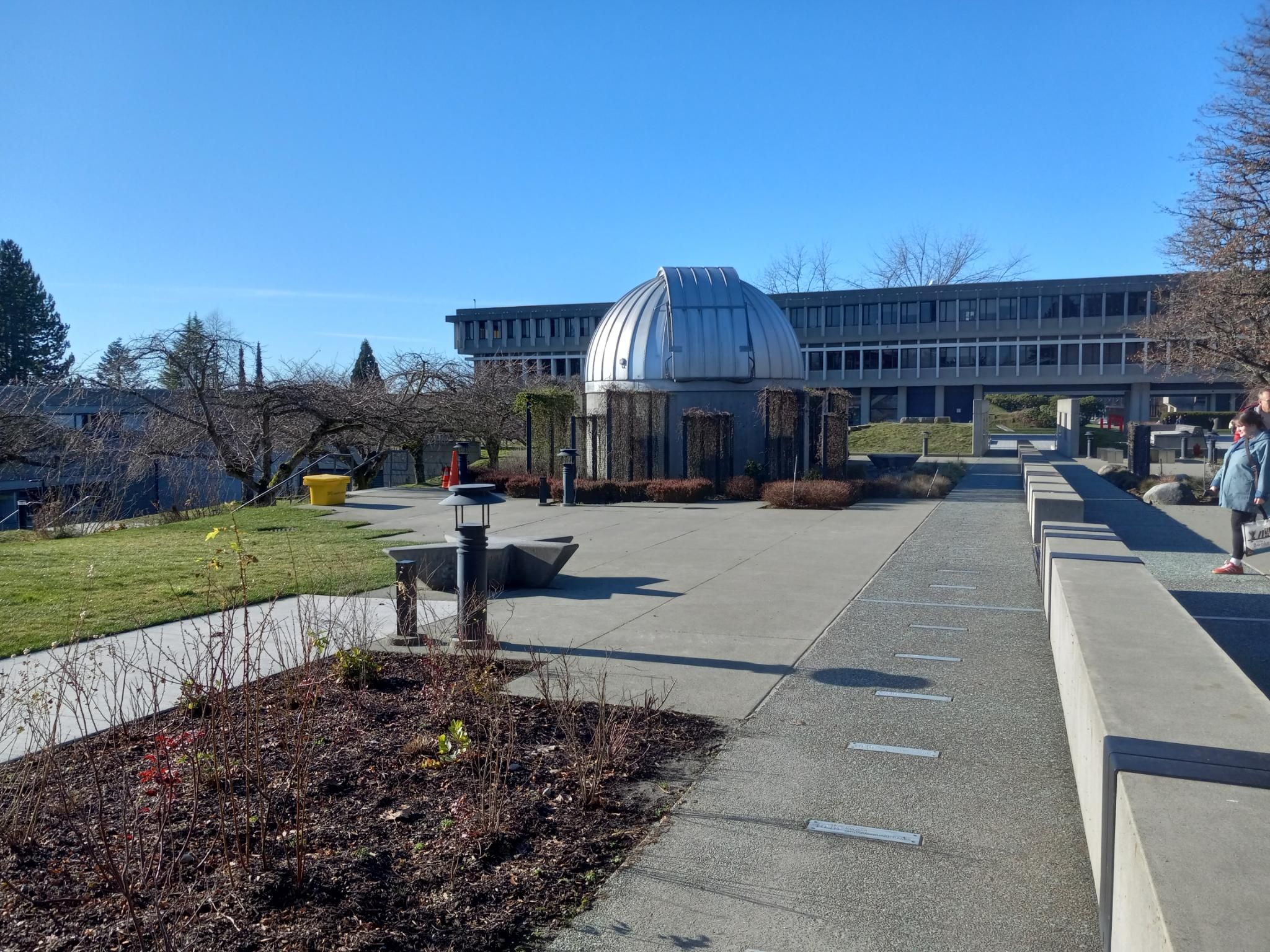 A telescope dome sitting in a courtyard under the blue sky.  There's a powers-of-ten art exhibit on the ground, a spectral lines art project on the concrete benches, and a bunch of star maps on some concrete walls further on.