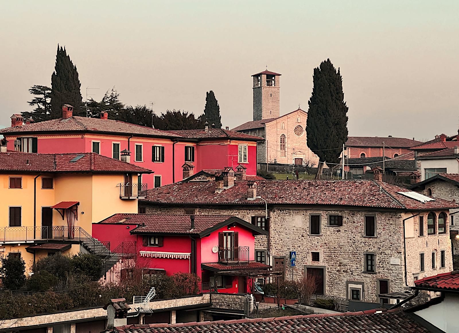 Paesaggio, tardo pomeriggio invernale, borgo antico, case in pietra ed intonacate di color rosa/fucsia, si vede il campanile di un convento, in pietra, vicino ad un alto cipresso sullo sfondo. 