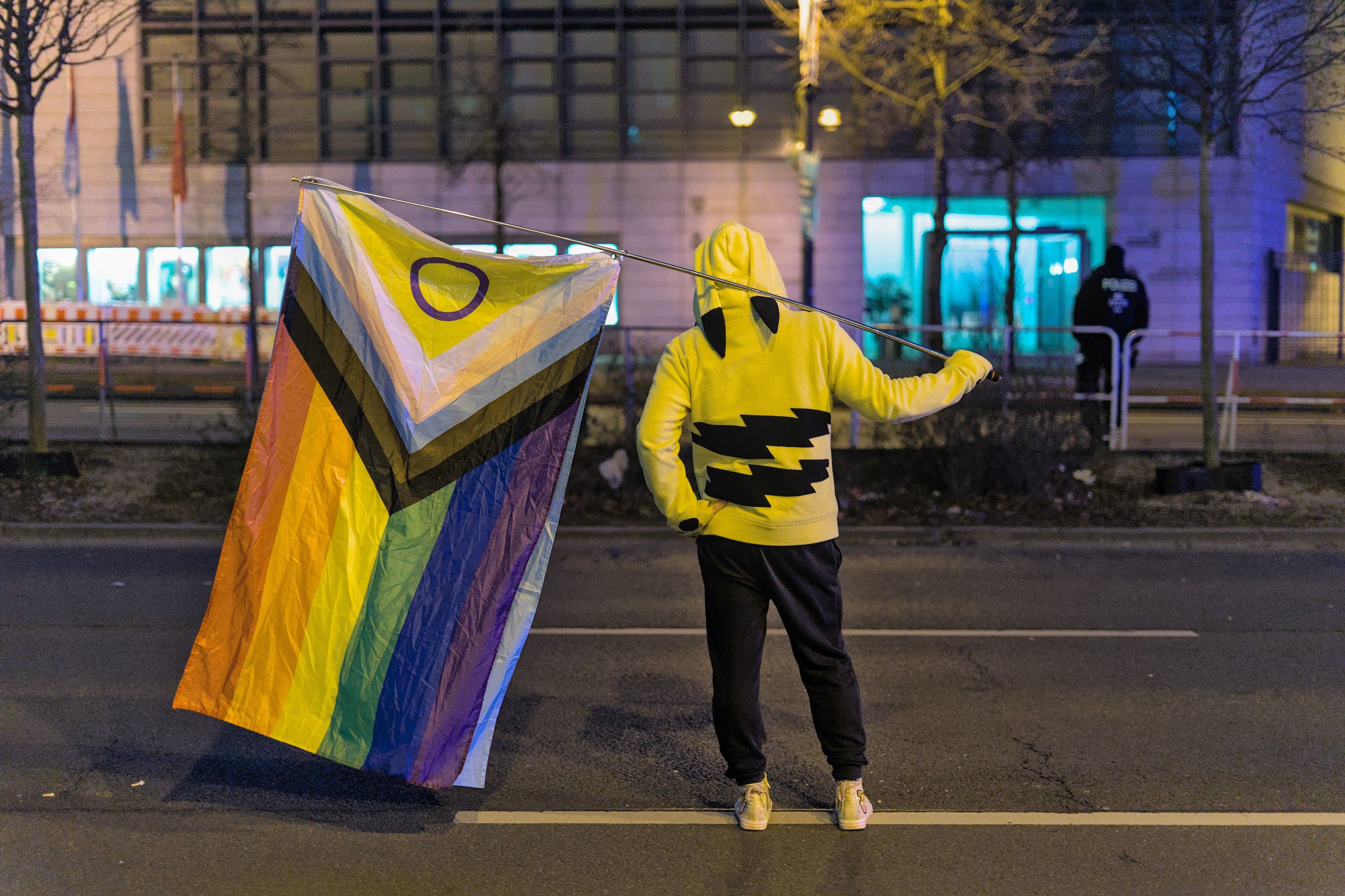 A (night-time) photo of someone standing on a road, wearing a Pikachu Hoodie while holding a progress pride flag with confident posture, back towards the camera. Quite in the background and out of focus, a police-person (german Polizei) is walking roughly away from the camera, relaxed posture. Next to that police-person, a building is illuminated with the telltale blue light of a police siren.
There is lots of colour and contrasts in this picture, but somehow the person standing in the center keeps pulling the focus to themself again.
The police was there out of routine with protests in Germany, clearing the roads of for the march. The photo was, of course, taken and published with permission.