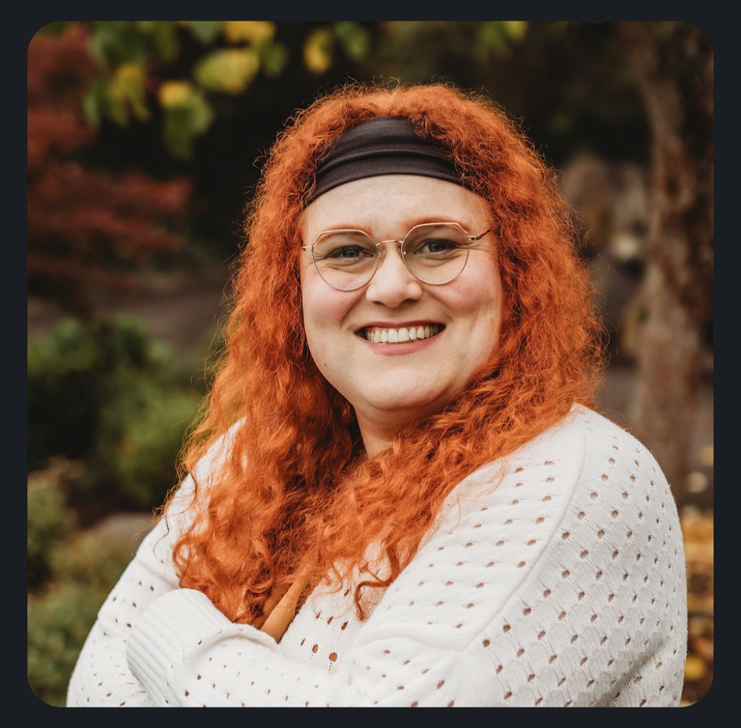 Headshot style photo of me, a white transfem person. Standing with my arms crossed, wearing an off-white sweater, glasses, and a black headband at my hairline. Curly copper hair rests below my shoulders on my chest. The background is autumn foliage.