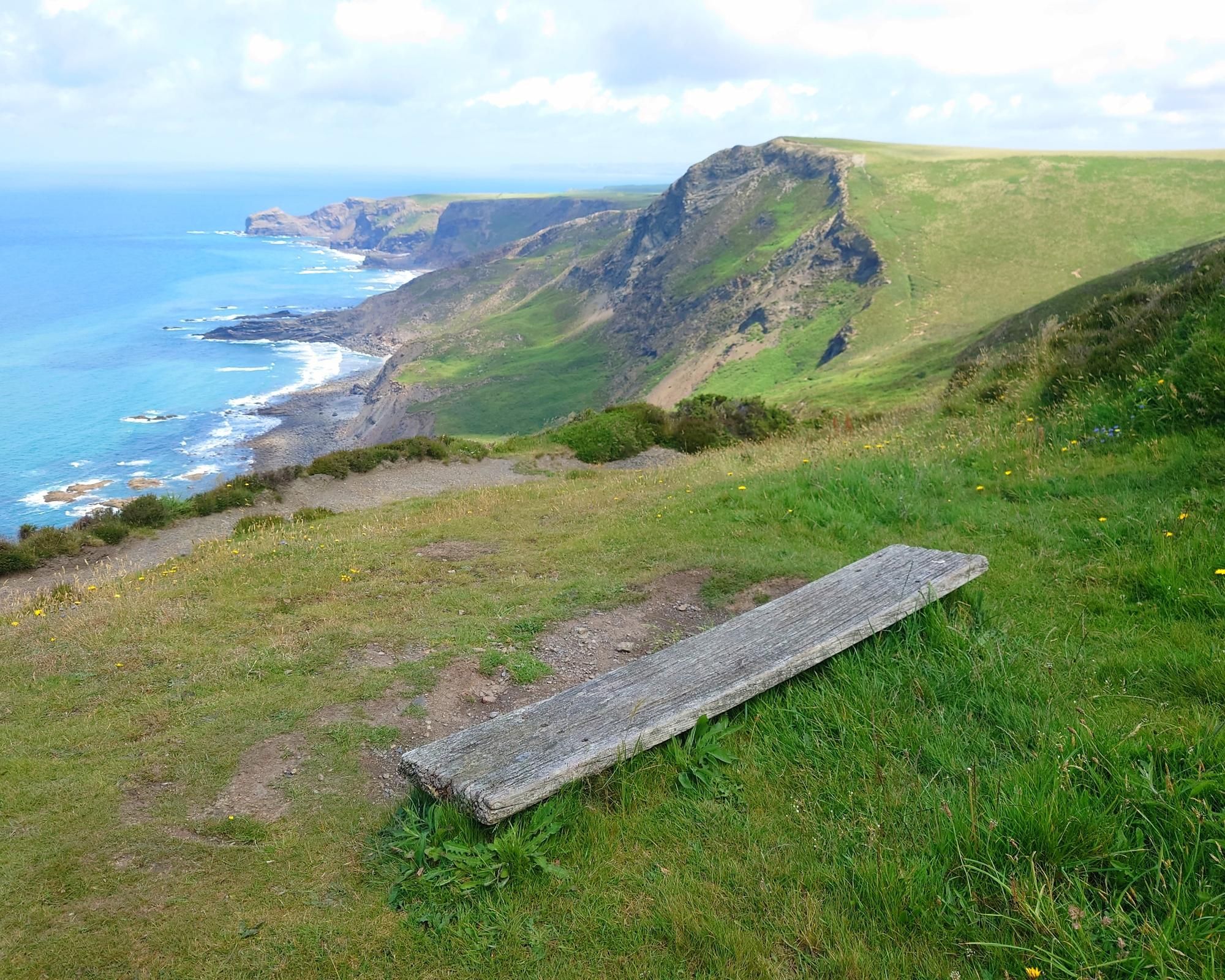 A seascape of blue ocean, slightly greying cumulus above, and headlands with green vegetation on the cliffs  and fields on top stretching off on the right hand side, with the foreground showing a simple weathered and low backless bench nearest the camera, which is angled towards sitting and taking in the view beyond.