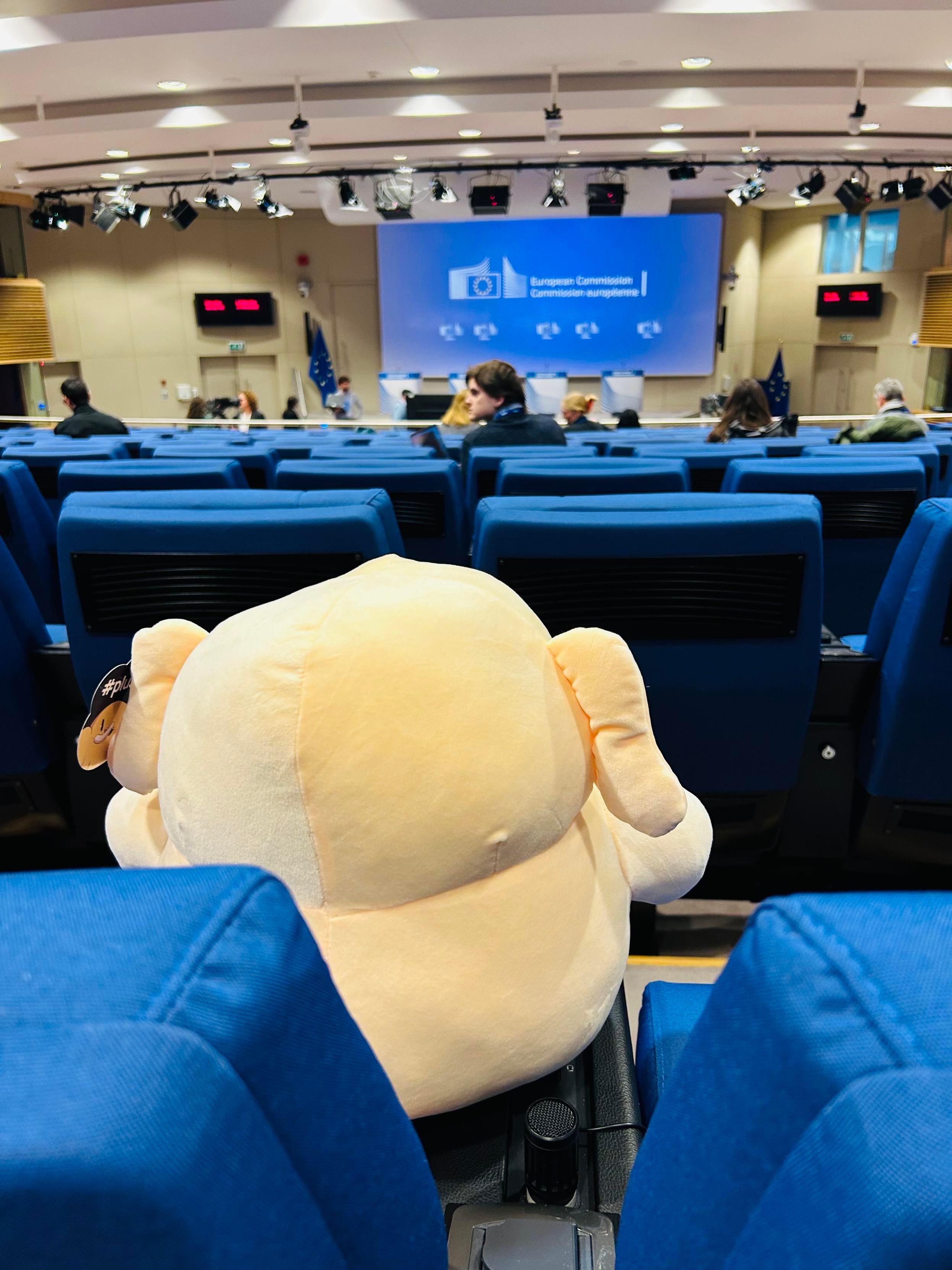 Mastodon plushie photographed from behind, hes sitting in the press room, in front of him are a couple of journalists and a screen featuring the European Commission logo and some podiums 