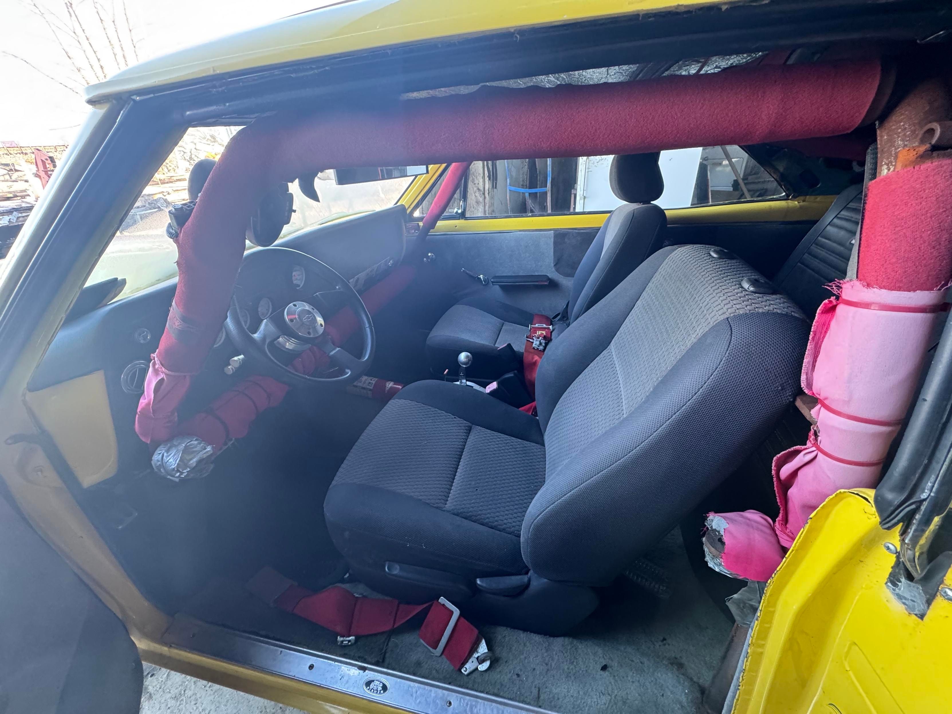 Interior of a yellow car featuring two front seats, a steering wheel with a custom steering wheel cover, and safety harnesses. The vehicle has visible roll cage padding, indicating modifications for racing or safety purposes.