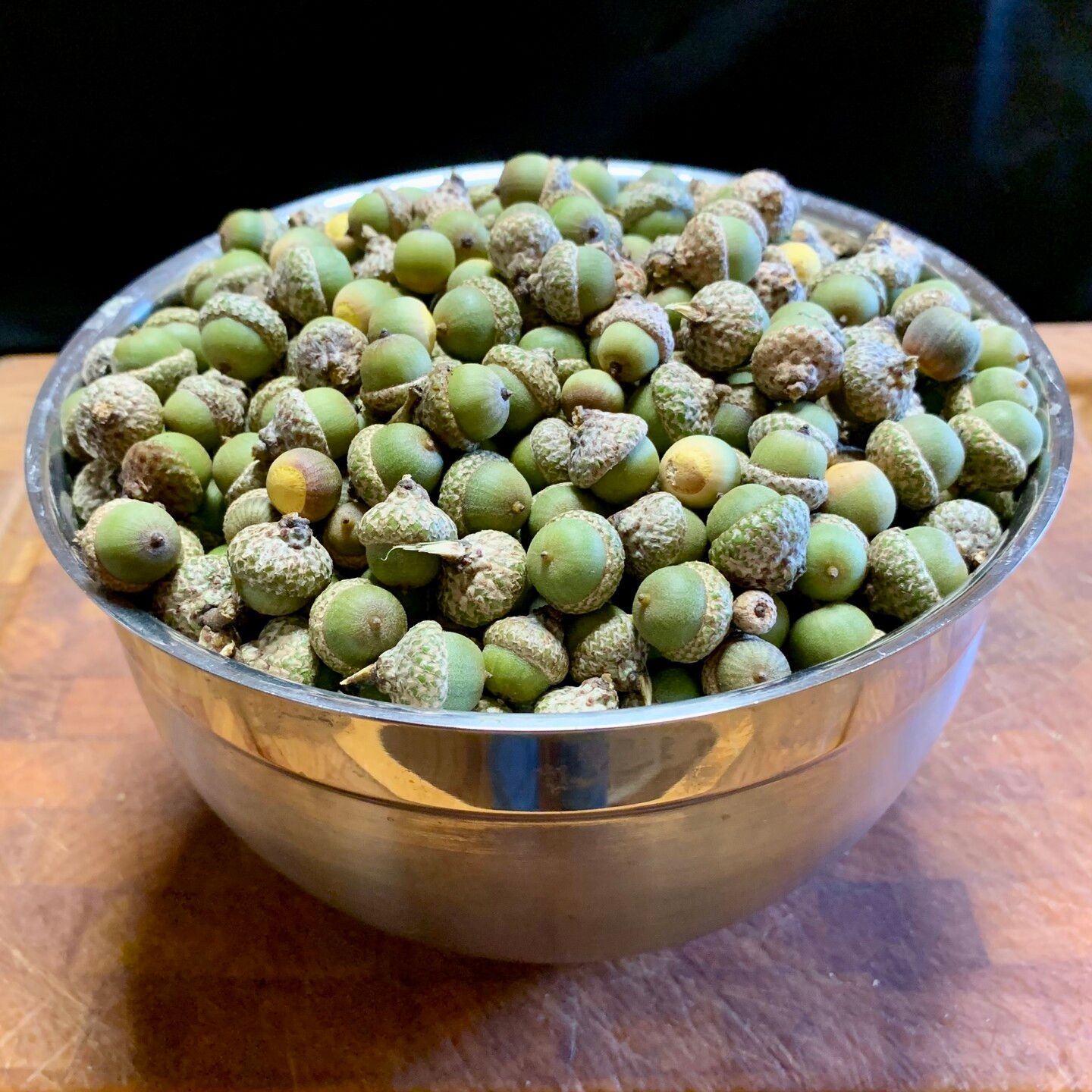 Black background, sitting on a sturdy brown woodblock is a silver bowl filled to the brim with pea-green little acorns. Some still have their grey caps while others have fallen off to reveal bright sunflower-yellow tops. So cheery! 
