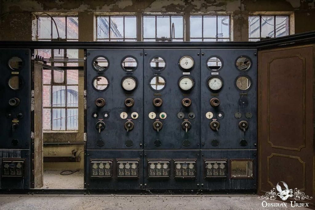 This image shows an old, dusty control panel with gauges, switches, and levers, suggesting an abandoned industrial setting. Sunlight streams through grimy windows, illuminating the decay. 