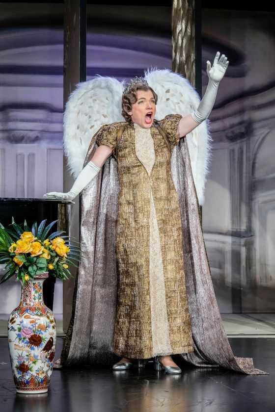 Eine Frau steht mit weit geöffnetem Mund singend auf einer Bühne. Die rechte Hand ruht auf einem Konzertflügel, der linke Arm ist theatralisch nach oben gestreckt. Sie trägt ein goldfarbenes Kostüm mit zwei Engelsflügeln am Rücken.