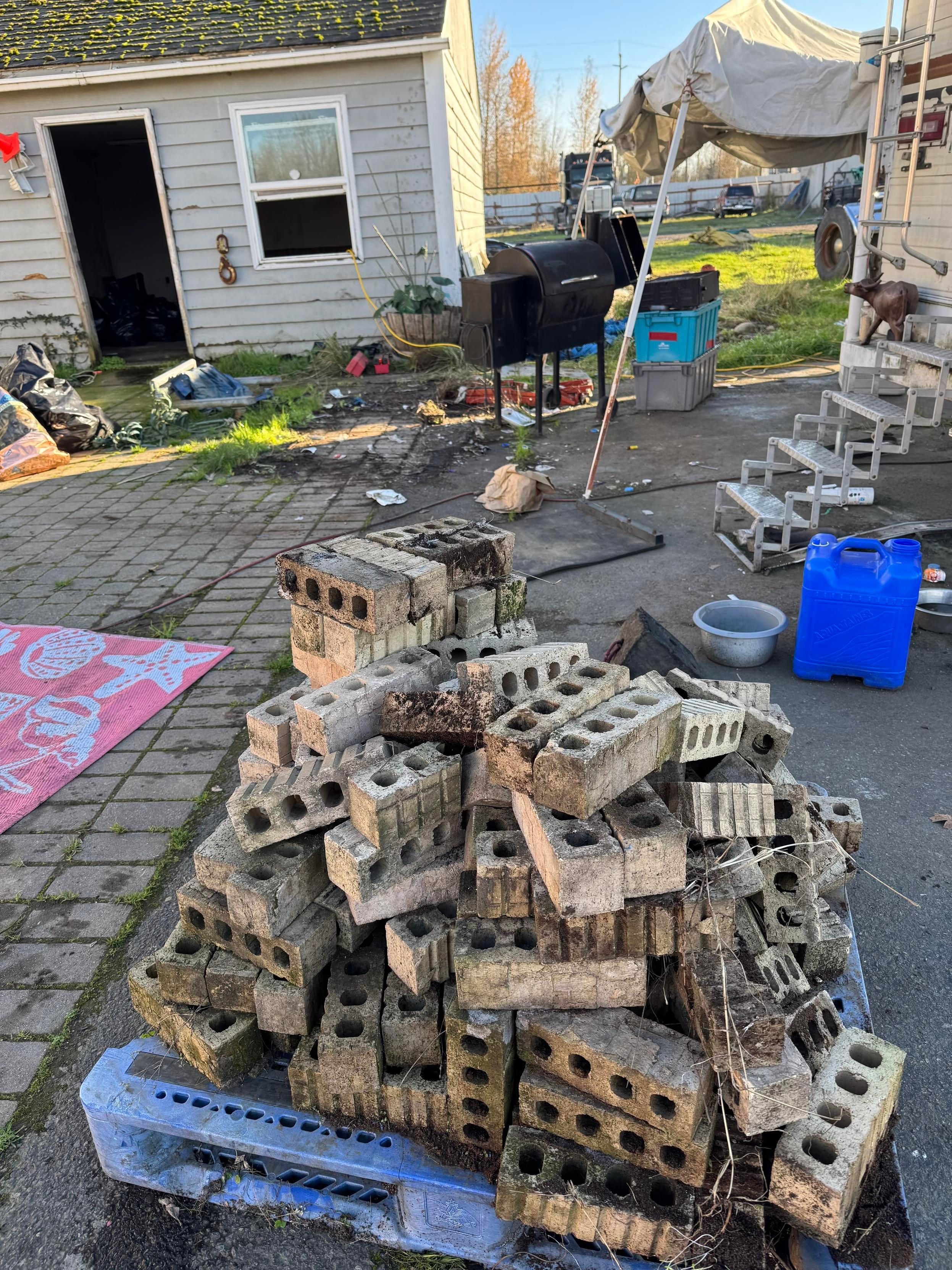 A pile of concrete blocks is stacked on a blue pallet, with a messy outdoor area in the background. Visible are a rundown house, a grill, various trash and debris, and some outdoor equipment. The scene appears cluttered and unkempt.