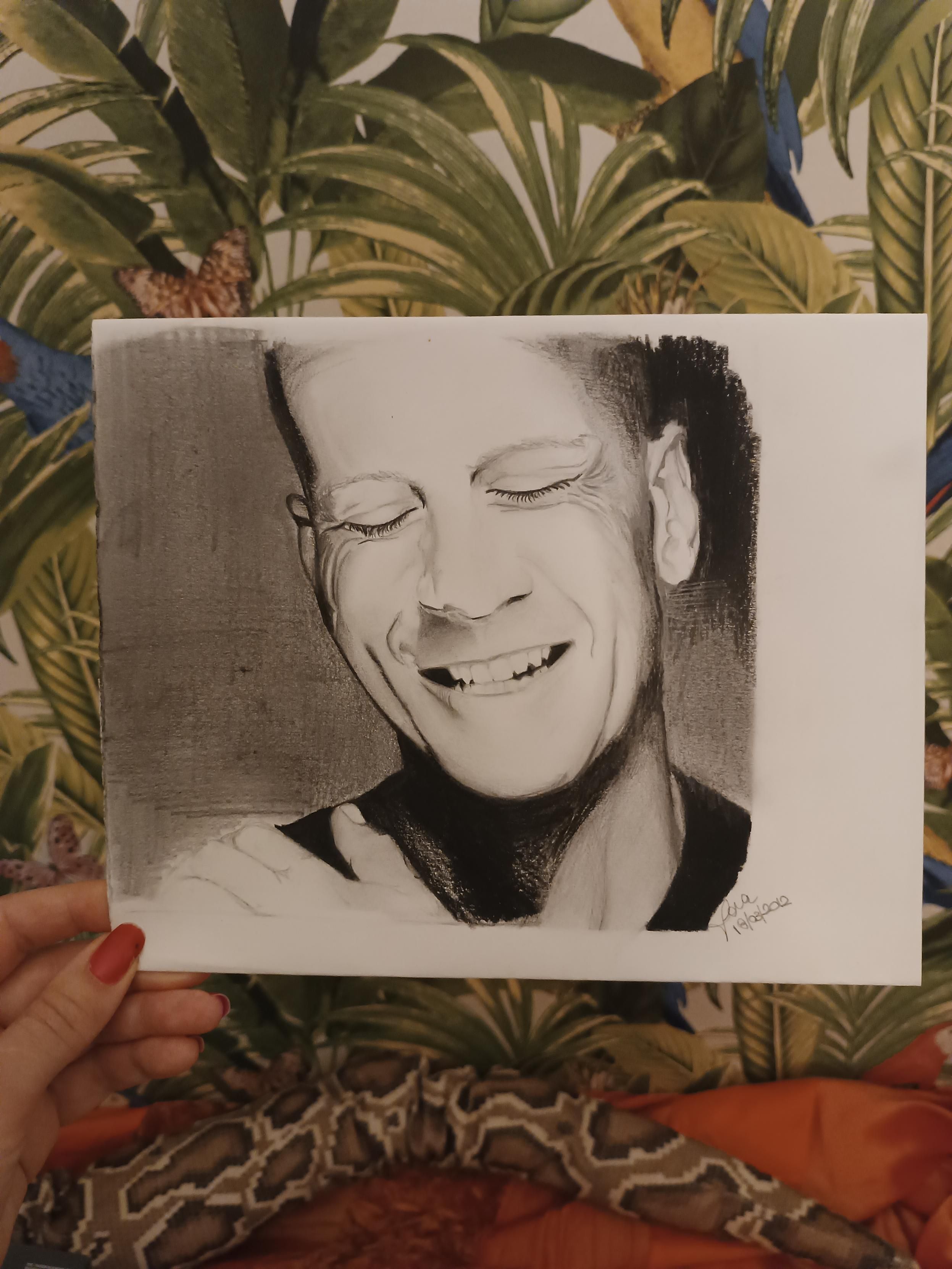 Ritratto in chiaroscuro di Bruce Willis sorridente, la mano tiene il foglio in un angolo, su sfondo carta da parati con foglie e pappagalli. Risale al 2012