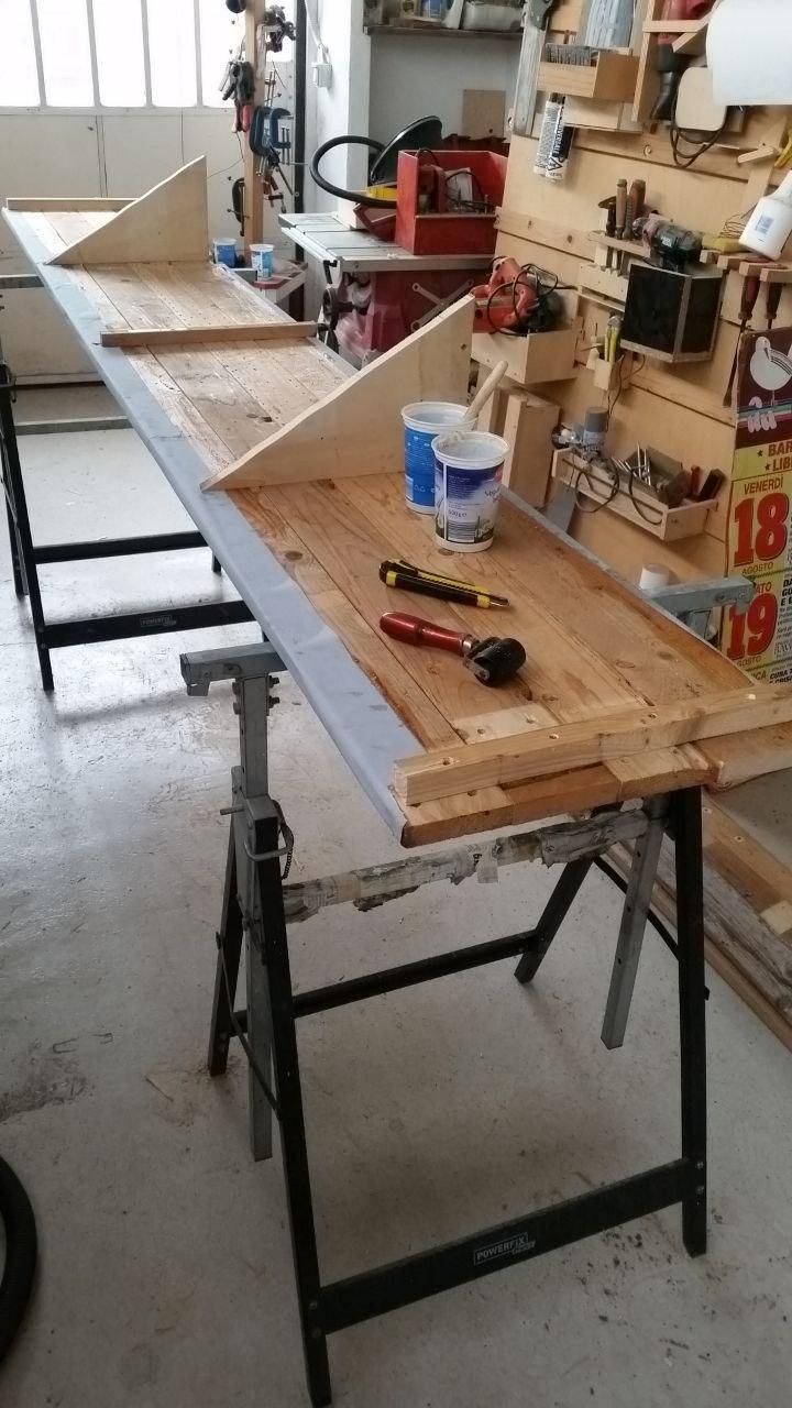 Piano dello scaffale capovolto.
Sui bordi si notano i manifesti risvoltati.
Le due "pinne" servono per fissarlo al supporto verticale.
Sul piano un cutter per rifilare i manifesti intorno alle sporgenze, un rullino per tappezzeria per spianare bene, un barattolo di yogurt (di recupero) pieno di acqua piovana (di recupero), con dentro un pennello da 2,5", e uno con la colla.
