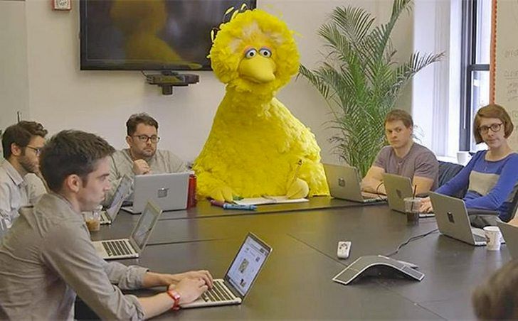 The image shows Big Bird from Sesame Street sitting at a long conference table with several people. Big Bird is in the center of the table, and the people around him are using laptops. It appears to be a meeting.