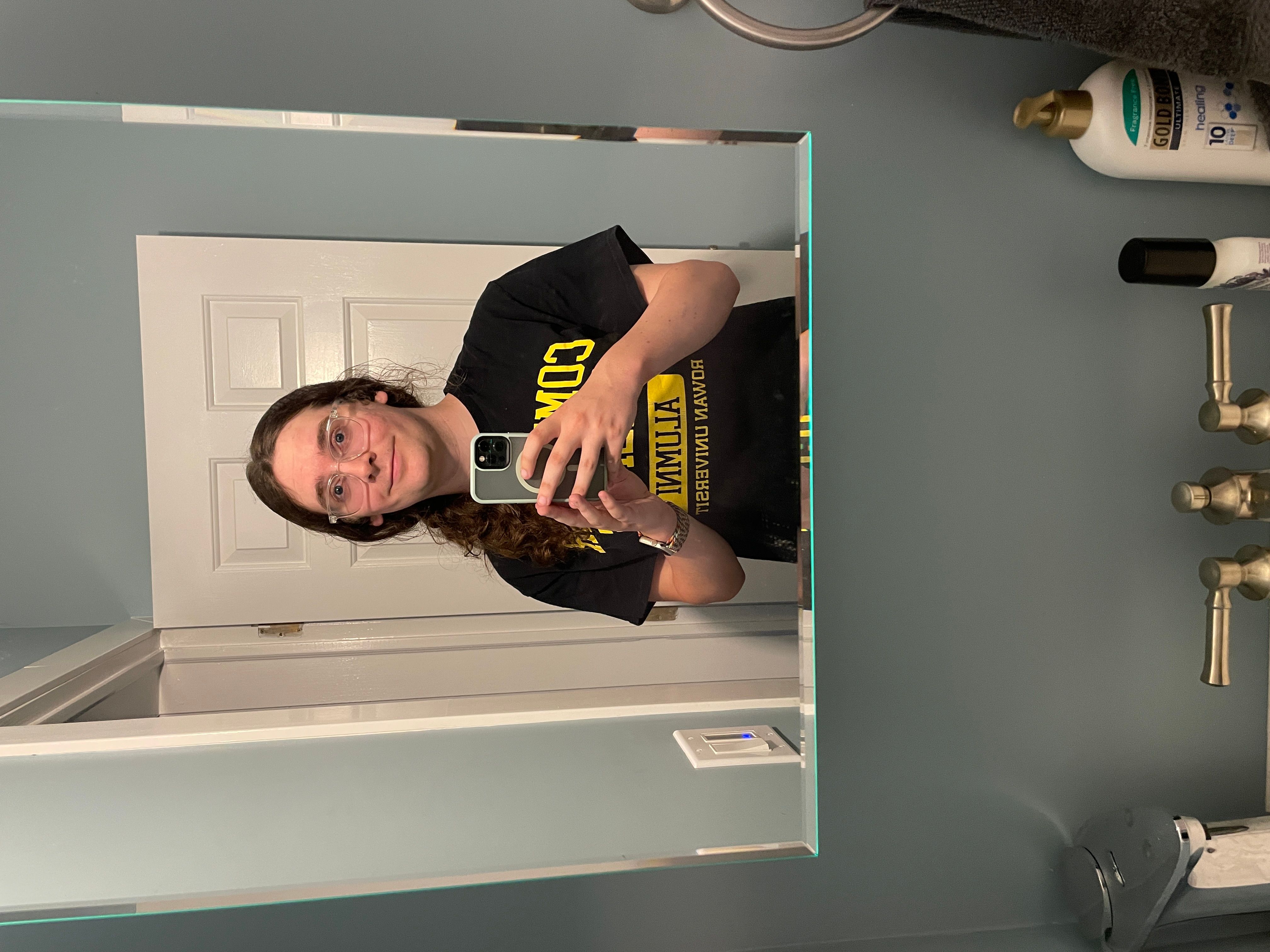 a white woman with dark brown curly hair is taking a selfie in a mirror. her hair is tied in a ponytail that is draped around her left shoulder. she is wearing clear-framed glasses and a black t-shirt with yellow lettering, the words being partially obscured by her arms holding a phone. she is smiling with her mouth closed and is looking up with wide eyes.