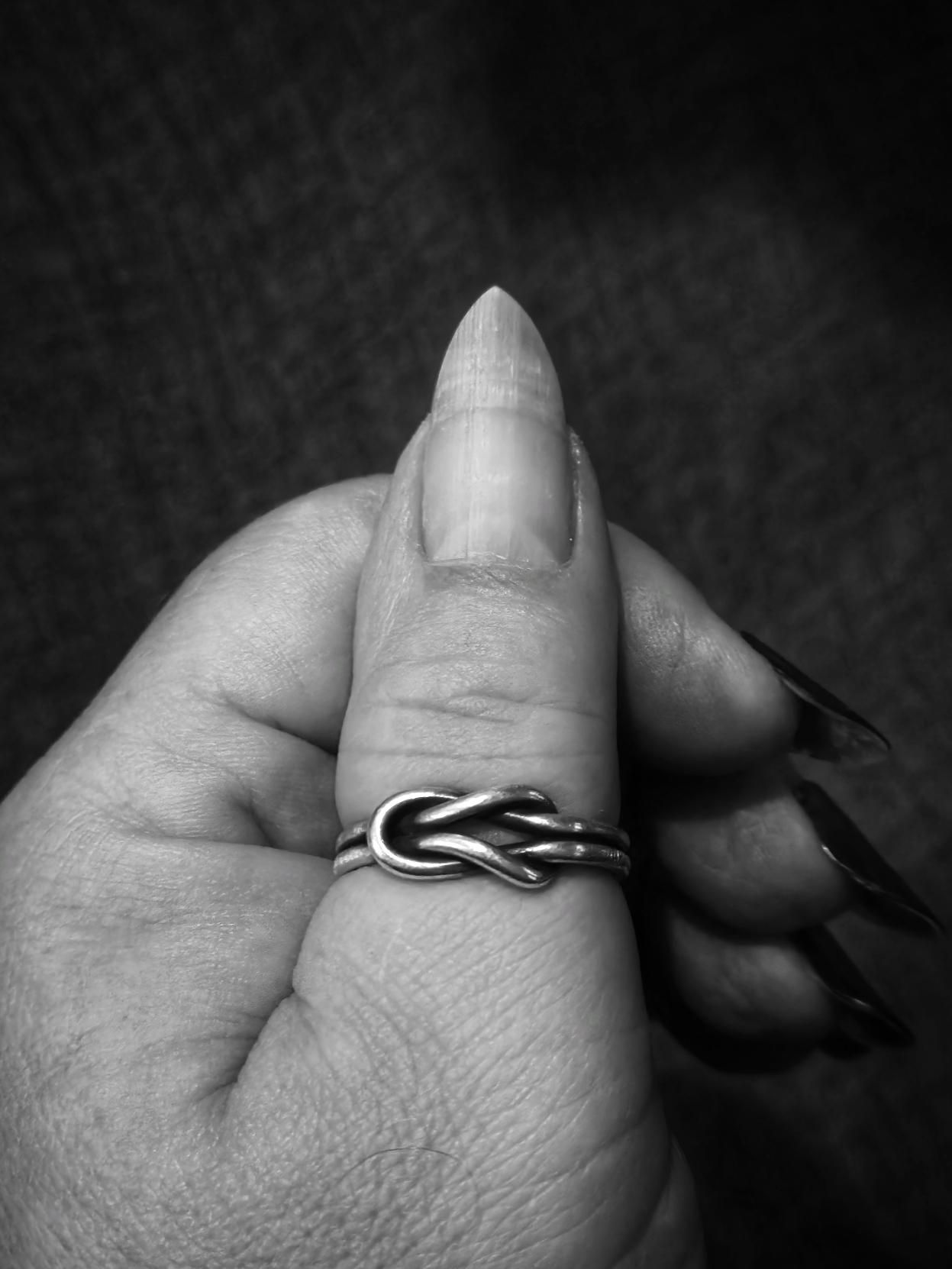 Close-up in black and white of a hand holding its own thumb upright, framed by long, dark nails curling in from behind. The thumbnail is shaped into a precise, hard point—clearly filed to a sharp tip rather than rounded. Its surface is smooth with a natural sheen, and faint vertical ridges run from the cuticle to the tip, catching the light in subtle highlights. The nail appears strong, intentional, and meticulously maintained. A double-banded diy silver ring with an elegant, interwoven knot design wraps tightly around the thumb. The background is dark and blurred, drawing full focus to the striking contrast of skin, polished nail, and intricate metal