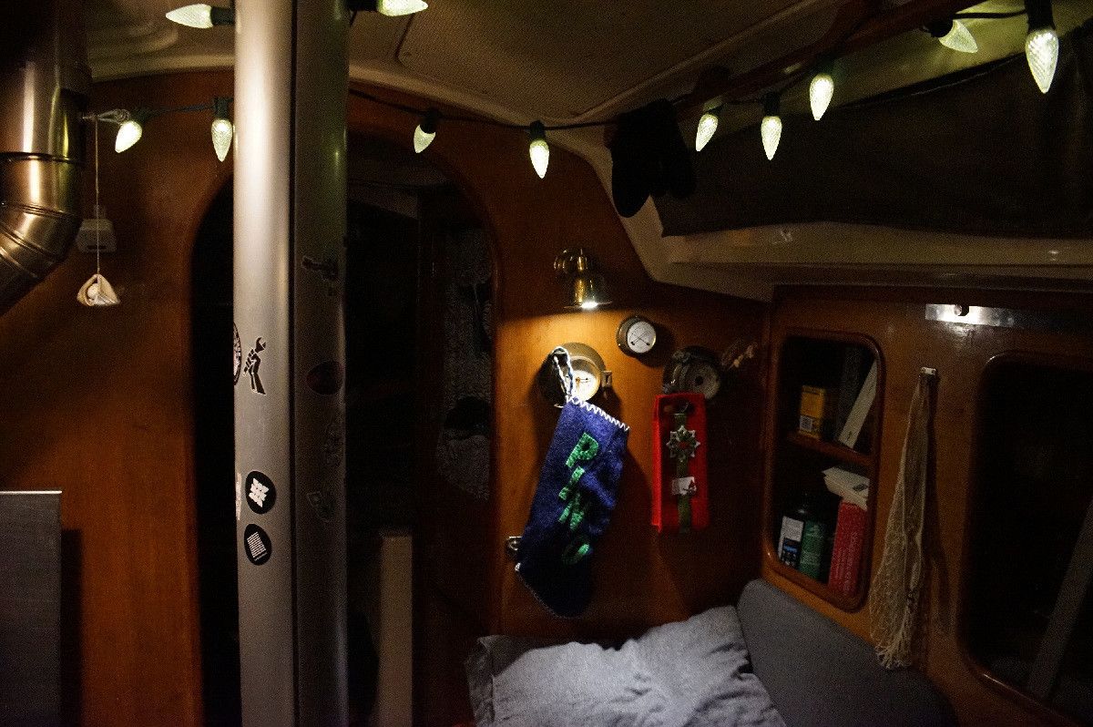 photo of the interior of a sailboat in late evening, decorated with a string of white lights, a blue felt christmas stocking with the name Pino on it, and a red felt bag. 