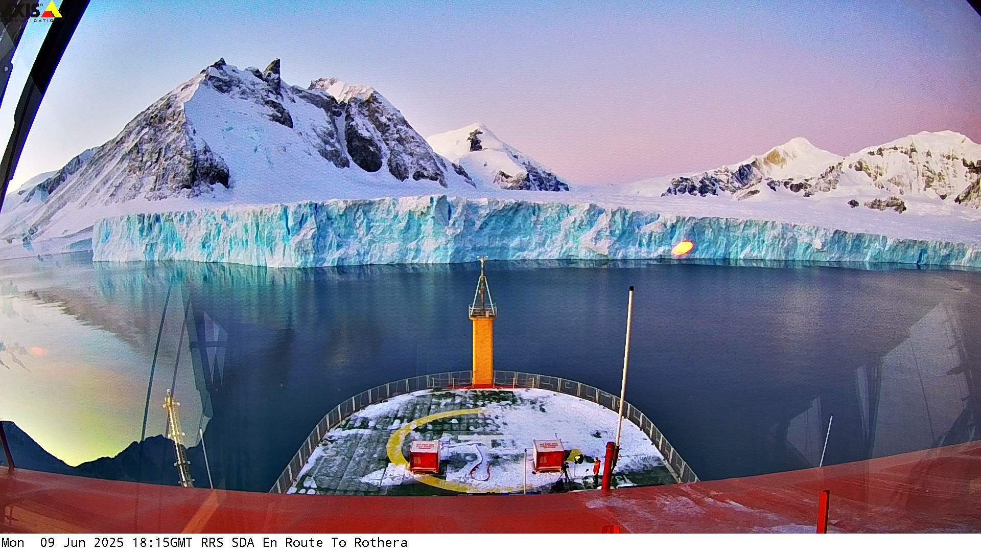A webcam photo captured by the RRS Sir David Attenborough as it stopped ahead of a frozen shore with snow-covered mountains in the distance. The colors are very vivid, with blues tinting the snow, contrasted with bright red and orange colors of the ship. The sky is a gradient of bright blue and pink.

Bottom of the image is captioned with the current date and time, and destination of the boat:

Monday June 9, 2025, 6:15pm, en route to Rothera.

(Note: Rothera is a name of a South Pole research station.)
