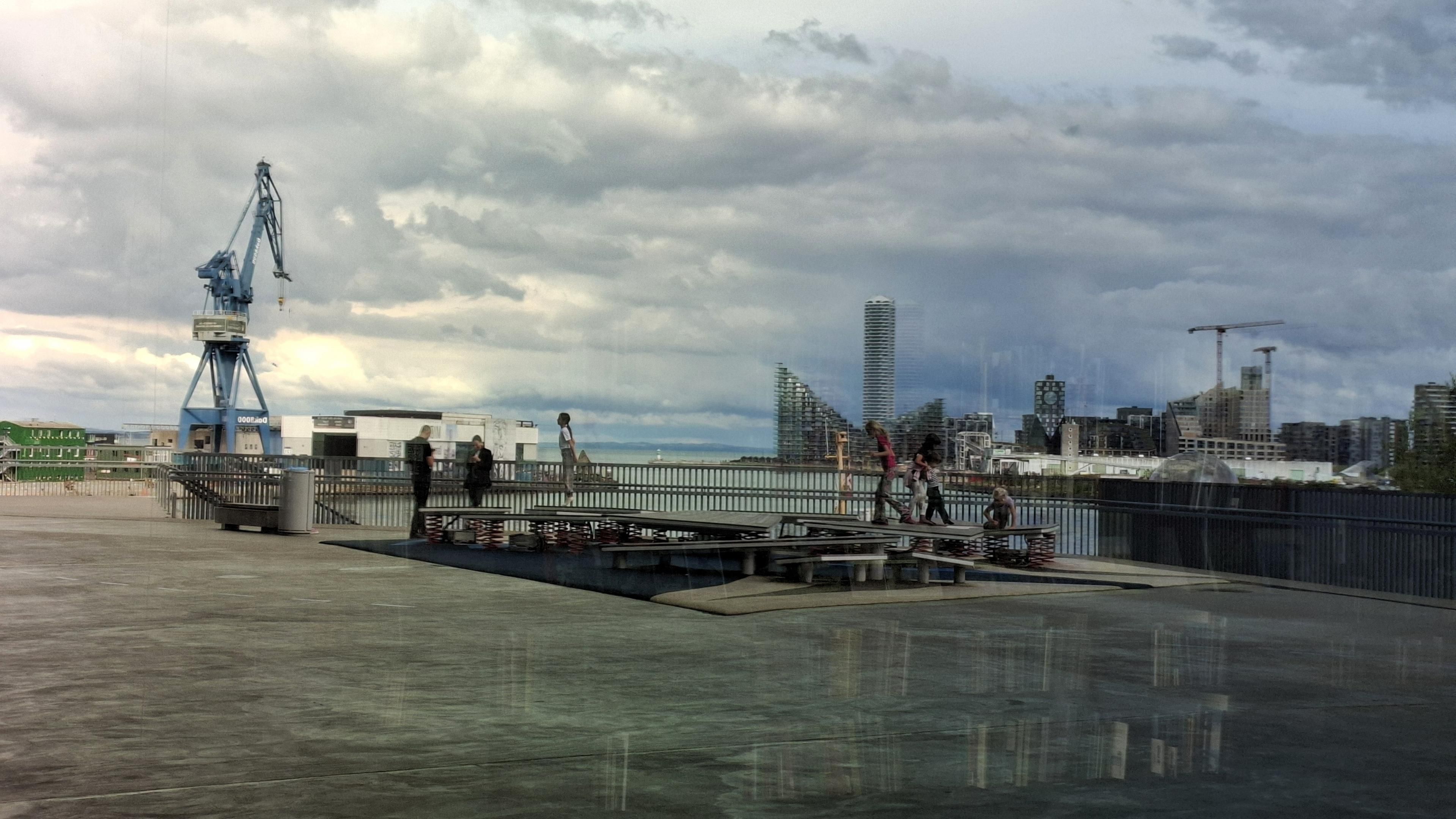 Short version: reflected in a window, a playground with harbour in background. Fuller description: looking in a huge picture window of Dokk1 Aarhus main library, Denmark, on an upper terrace. You can’t see the window edges, so it might not be obvious at first glance that this is a window. Inside, ghostly, indistinct in interior darkness, almost as if floating: rows of books on barely visible shelves. Overlaying them in the window, the full reflection of the scene behind us – the city and harbour. We can see a 75-metre tall industrial crane, angular like a blue  iron giraffe; Denmark’s tallest building, the narrow rectangular 142-metre tall Lighthouse apartment block; an A-shaped apartment block; a 70-metre high apartment block with a giant clock; calm light blue sea; overcast sky swirling with black and white clouds. On the terrace: playground platforms with indistinct figures of children walking and hopping and playing on them – one child is in mid-air, mid-hop.