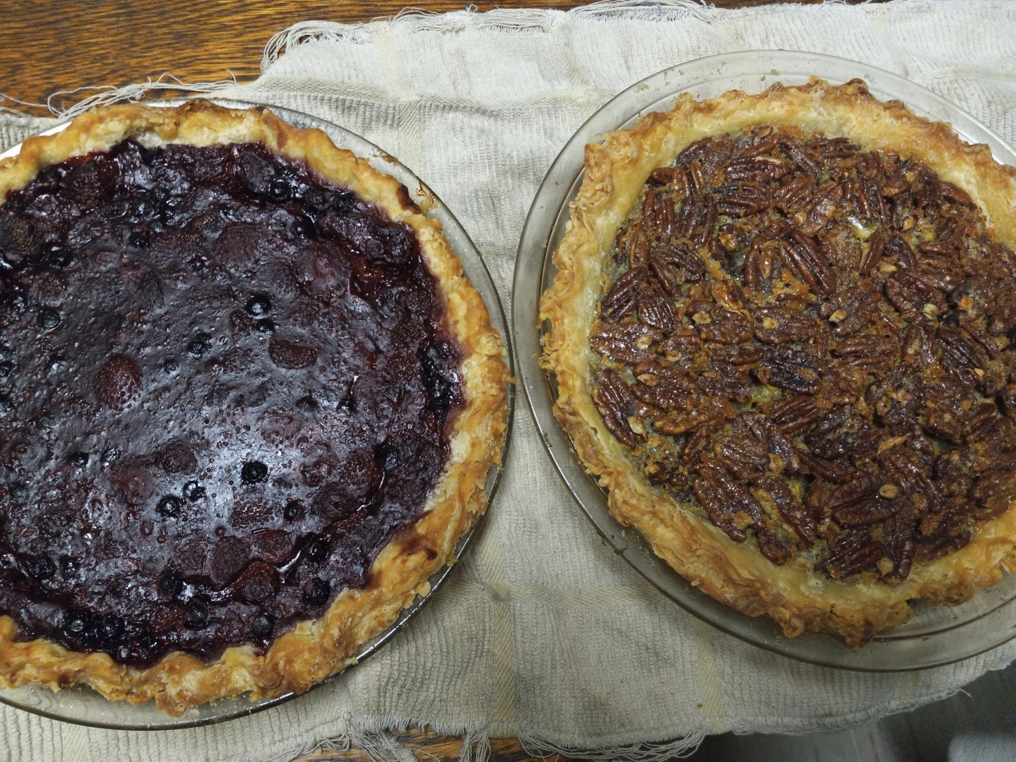 A berry pie on the left, and a pecan pie on the right, both in glass pie pans and sitting on an old towel while cooling.
