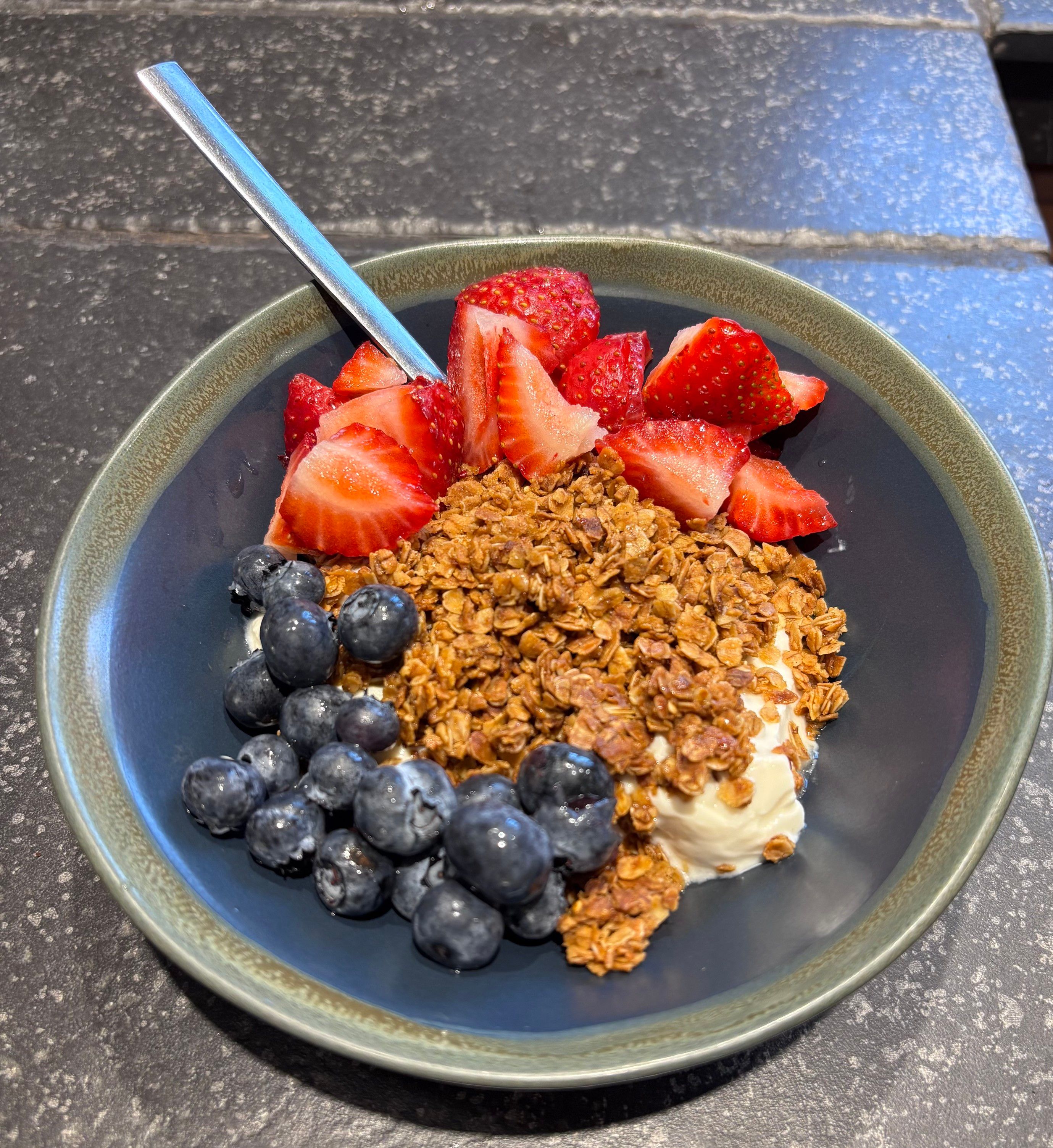 A bowl of yogurt topped with granola, sliced strawberries, and blueberries, with a spoon resting inside.
