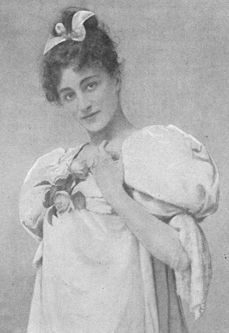 Edith Ostlere, a young white woman with dark curls in an updo, wearing a light colored dress with big puff sleeves