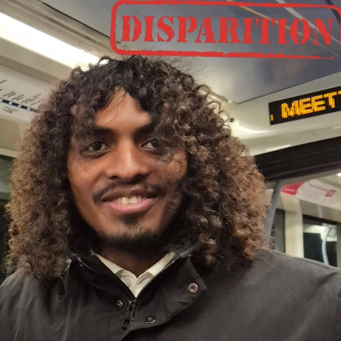 Smiling man with long, curly brown hair and a short beard, wearing a black jacket and a white collared shirt, inside a public transport vehicle with a red "DISPARITION" stamp above his head.