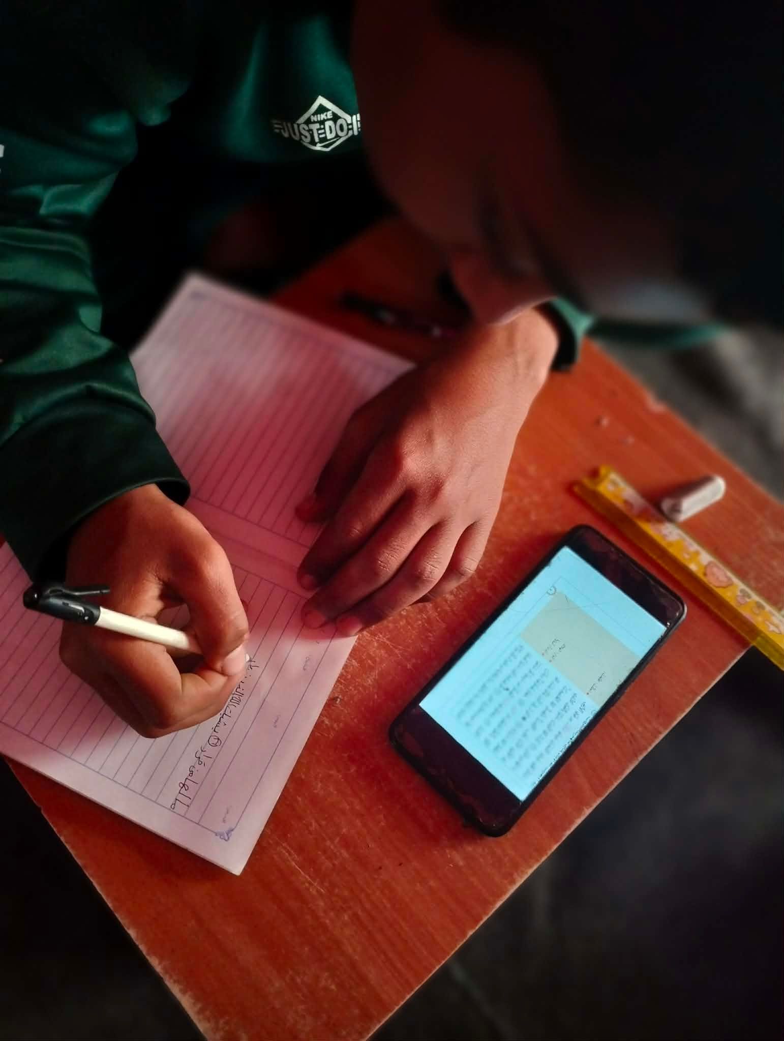 Abdul Rahman writing on a notebook with a pen on a wooden desk, with a smartphone displaying text and a ruler nearby. The person is wearing a green jacket.