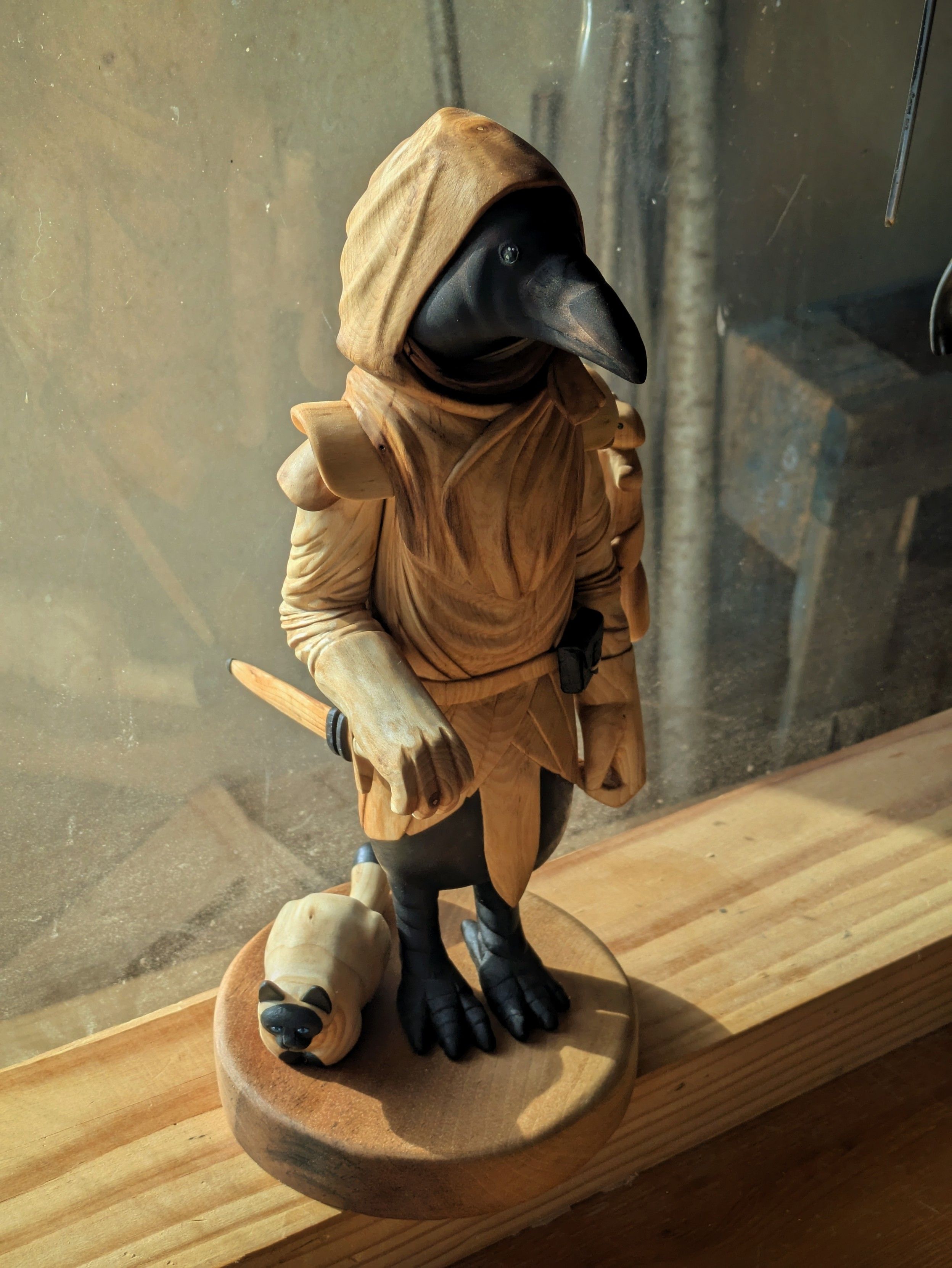 Woodcarved humanoid crow with a hooded cloak, a sword, satchel on its belt. It has a squirrel climbing its arm, and a cat sitting next to its feet