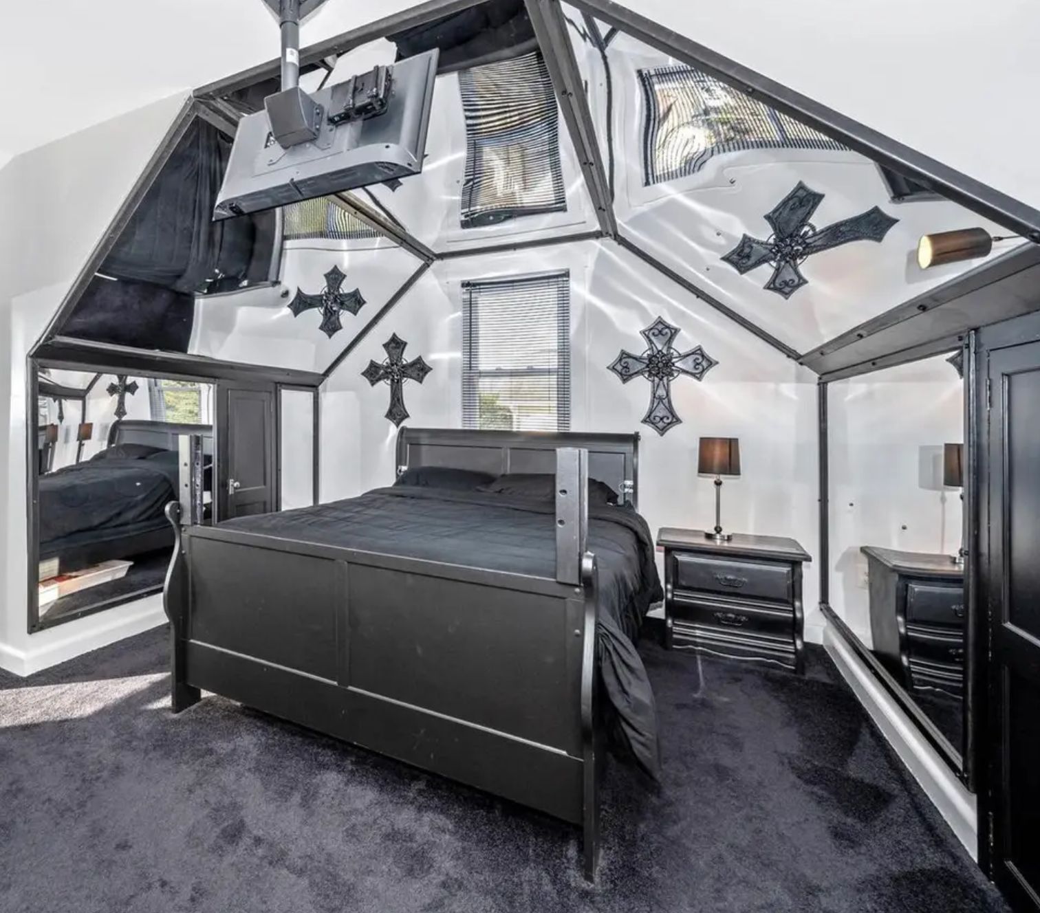 A bedroom with a black carpet and black bed with black linens. There are 2 goth style black crosses on the back wall. There is a vaulted ceiling with 3 mirrors above the bed, reflecting the crosses. There is a tv mounted on the ceiling at the foot of the bed