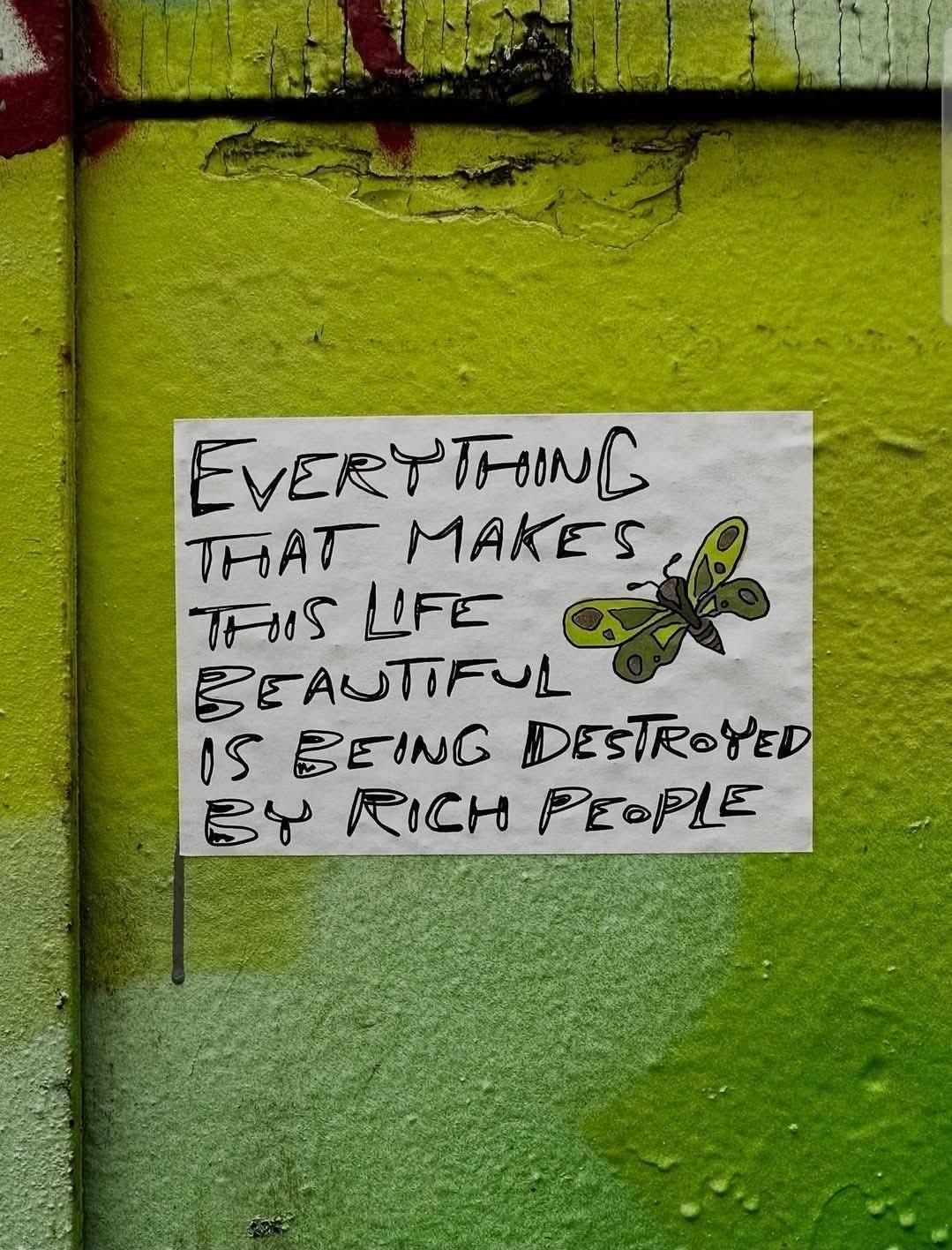 A handwritten poster on a textured green wall reads: “Everything that makes this life beautiful is being destroyed by rich people.” The text is written in uneven, expressive black letters, accompanied by a green and black butterfly illustration. The poster is taped or pasted to a lime-to-olive gradient background with faint graffiti marks and weathered paint, evoking a raw urban aesthetic and environmental message.