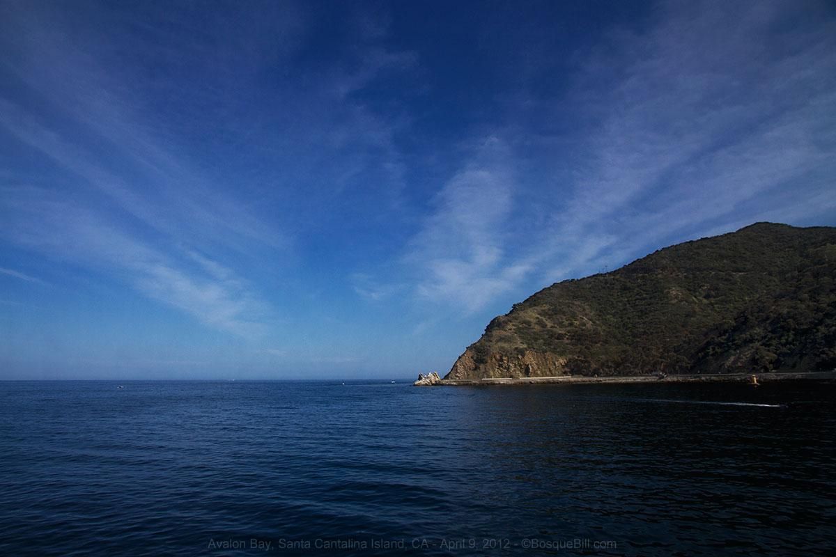 Calm very dark blue sea; dark blue sky with light clouds seemingly radiating from the center; a wedge of dark green land extends from right to the center.
©BosqueBill.com