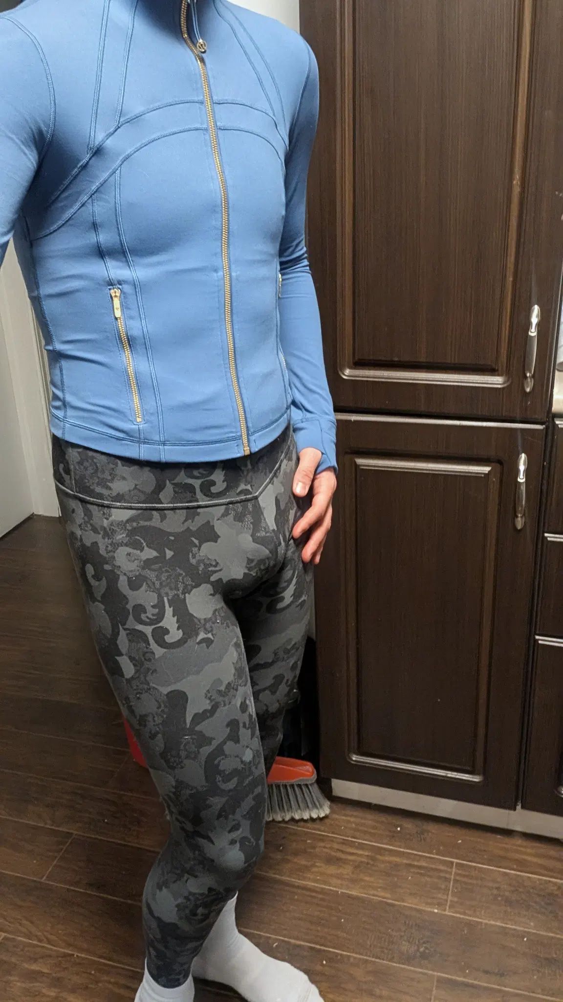 I stand in the kitchen with brown wooden cabinetry behind me. I am wearing black and grey leggings with a lace-like pattern. On top I'm wearing a blue, slim-fitting, full-zip, track-like jacket. The zipper is cold coloured with gold pocket zippers.
My hand rests on my hip, and the stitching on the jacket accentuates my form