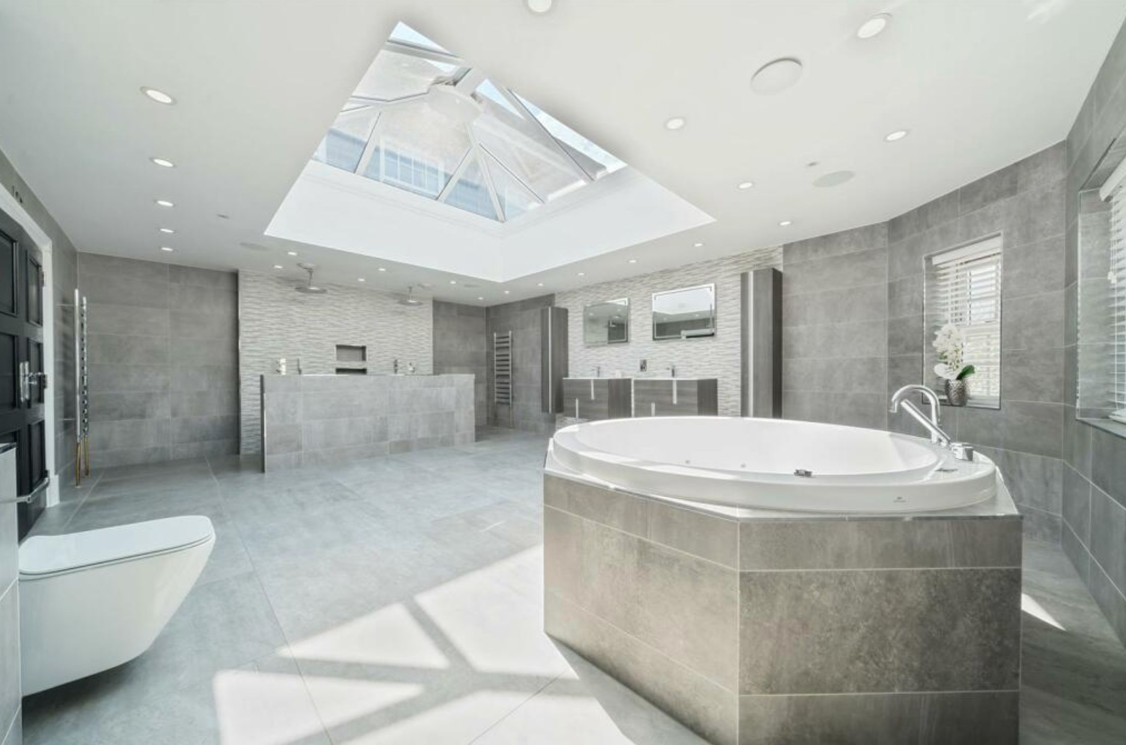A gigantic bathroom clad in grey tiles, with a round standalone bath, toilet, and shower at the far end. There is a lot of empty space and the overall effect is rather soulless. 