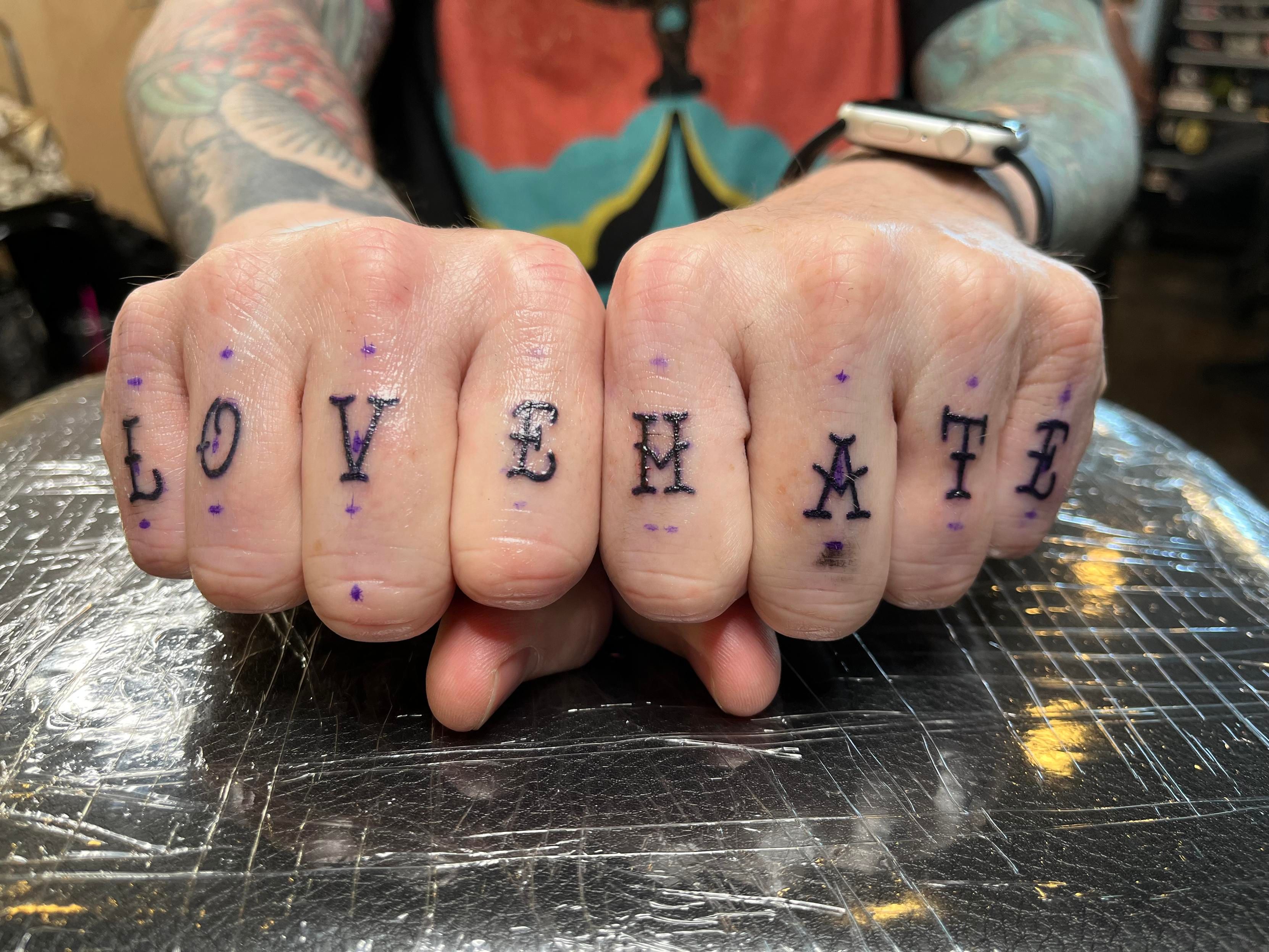 My hands in fists, resting close together on surface. My fingers show “love hate” freshly tattooed on them in a retro tattoo style typeface. 