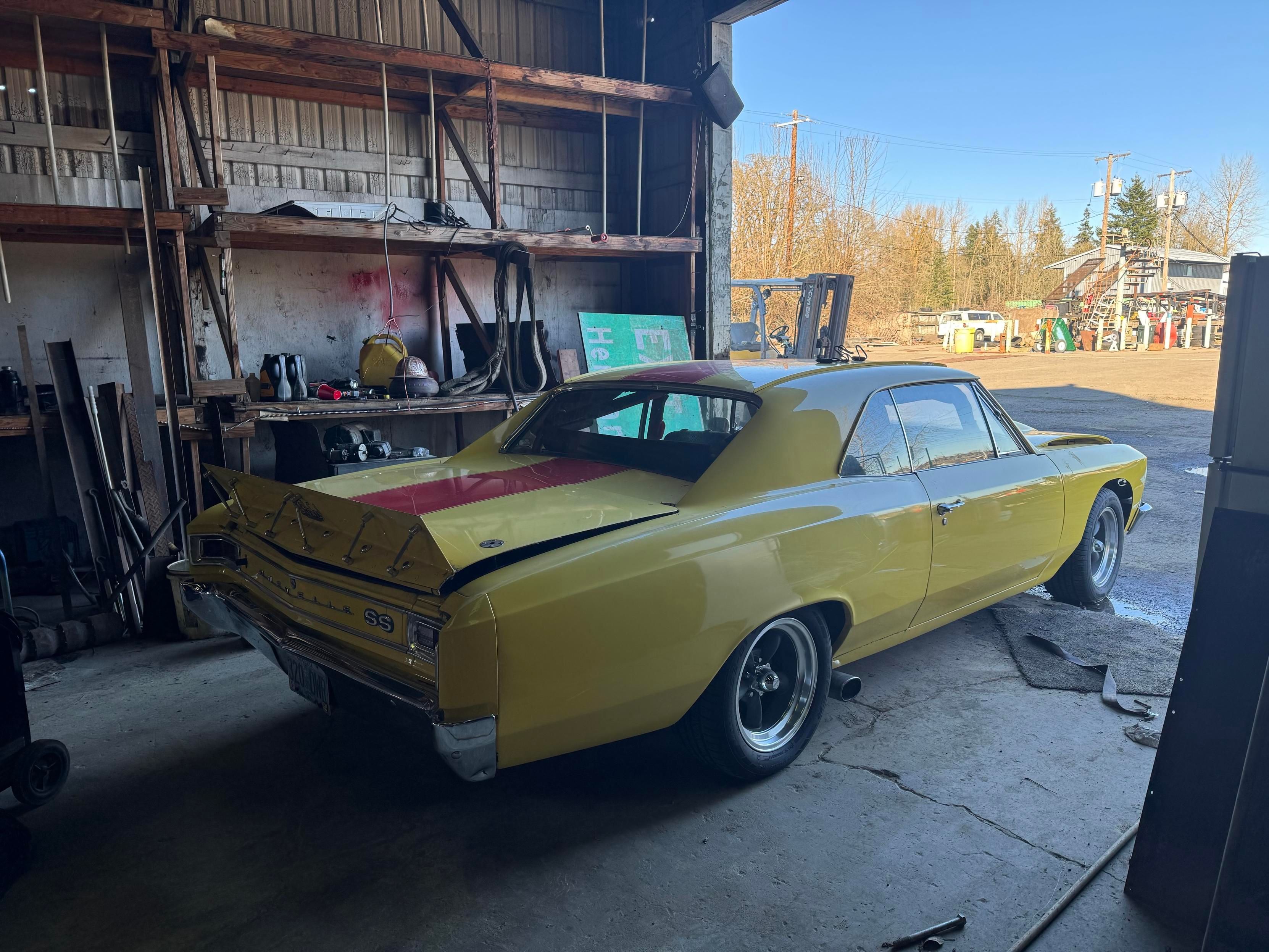 A yellow vintage car, prominently featuring a rear spoiler, is parked inside a garage. The area includes shelves with tools and equipment, and the door leads to an outdoor view with trees and additional vehicles.