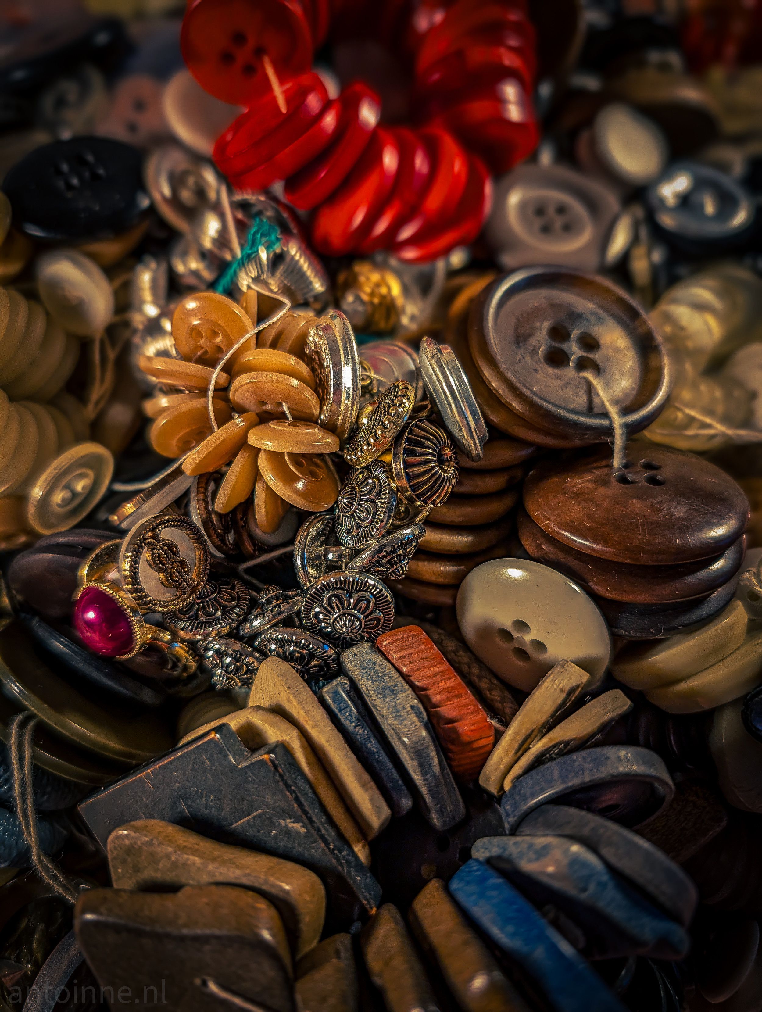 A rich, close-up photograph of a dense and eclectic collection of antique and vintage buttons. The composition is chaotic yet aesthetically pleasing, filled with a vast variety of shapes, colors, and textures.

The overall palette is warm and earthy, dominated by muted browns, tans, and greys. However, several pops of color break through the neutral tones.

The collection showcases a diverse range of materials. There are buttons made of wood, horn, bone and shell. Several intricate, silver-toned metal buttons feature embossed floral or geometric patterns, adding a sense of craftsmanship and detail.