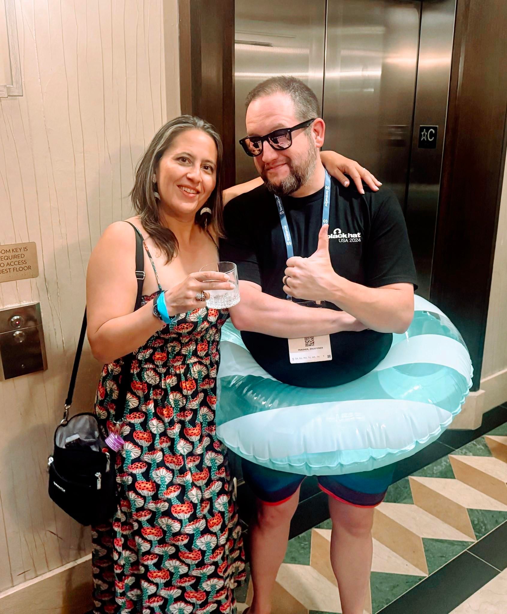 Photograph of a man and a woman in the Mandalay Bay hotel near the elevators to upper floors.  She is holding a cocktail and wearing a pretty dress and has her arm around the man who is wearing rainbow striped bathing trunks, pink sunglasses, and inexplicably has a large pool floatie around his hips.  The man is naturally giving a thumbs up with a grin.