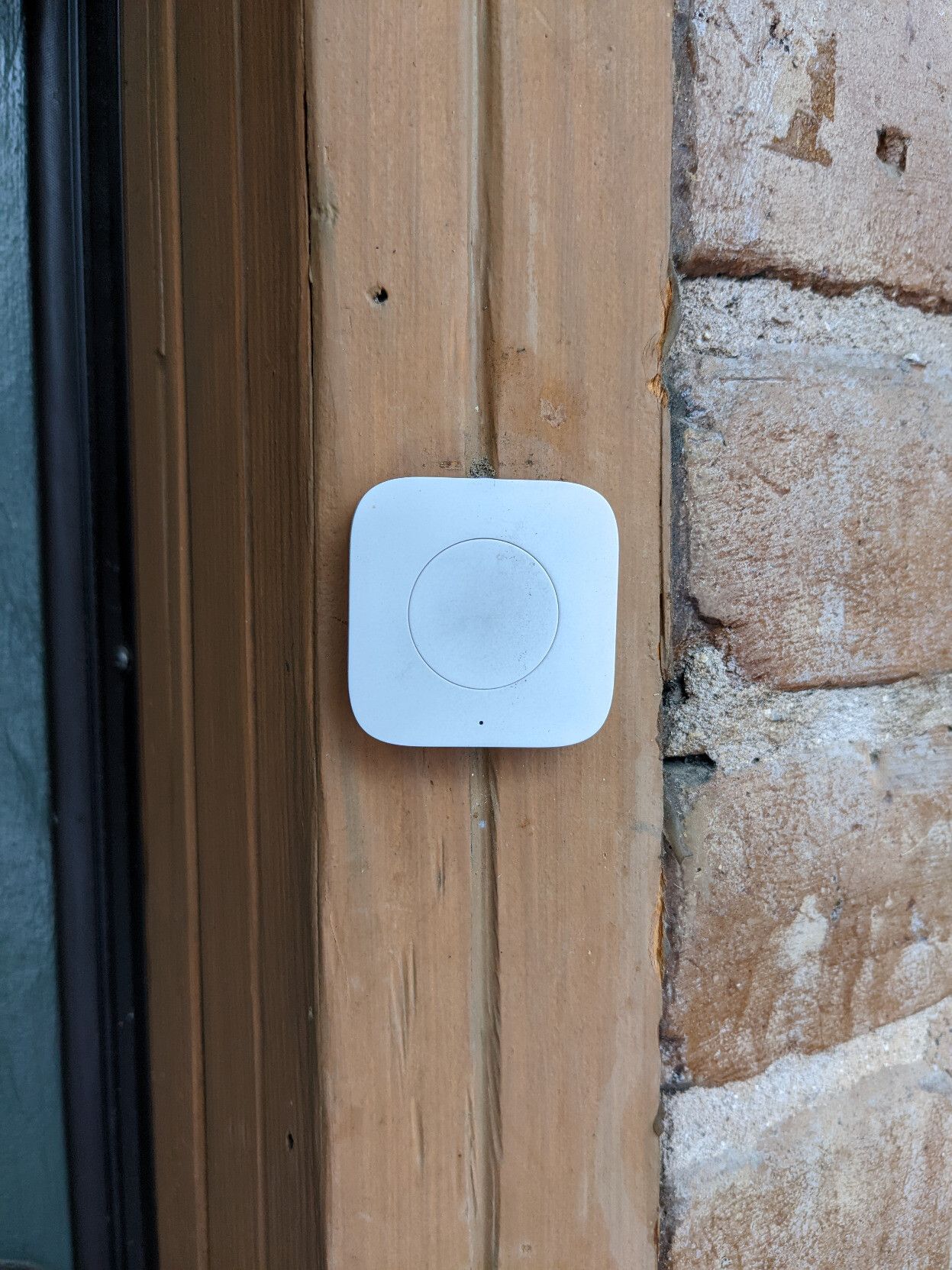 A nondescript round white button (a doorbell) mounted on a vertical wood member. To the left a part of a door is visible, and to the right, bricks.
