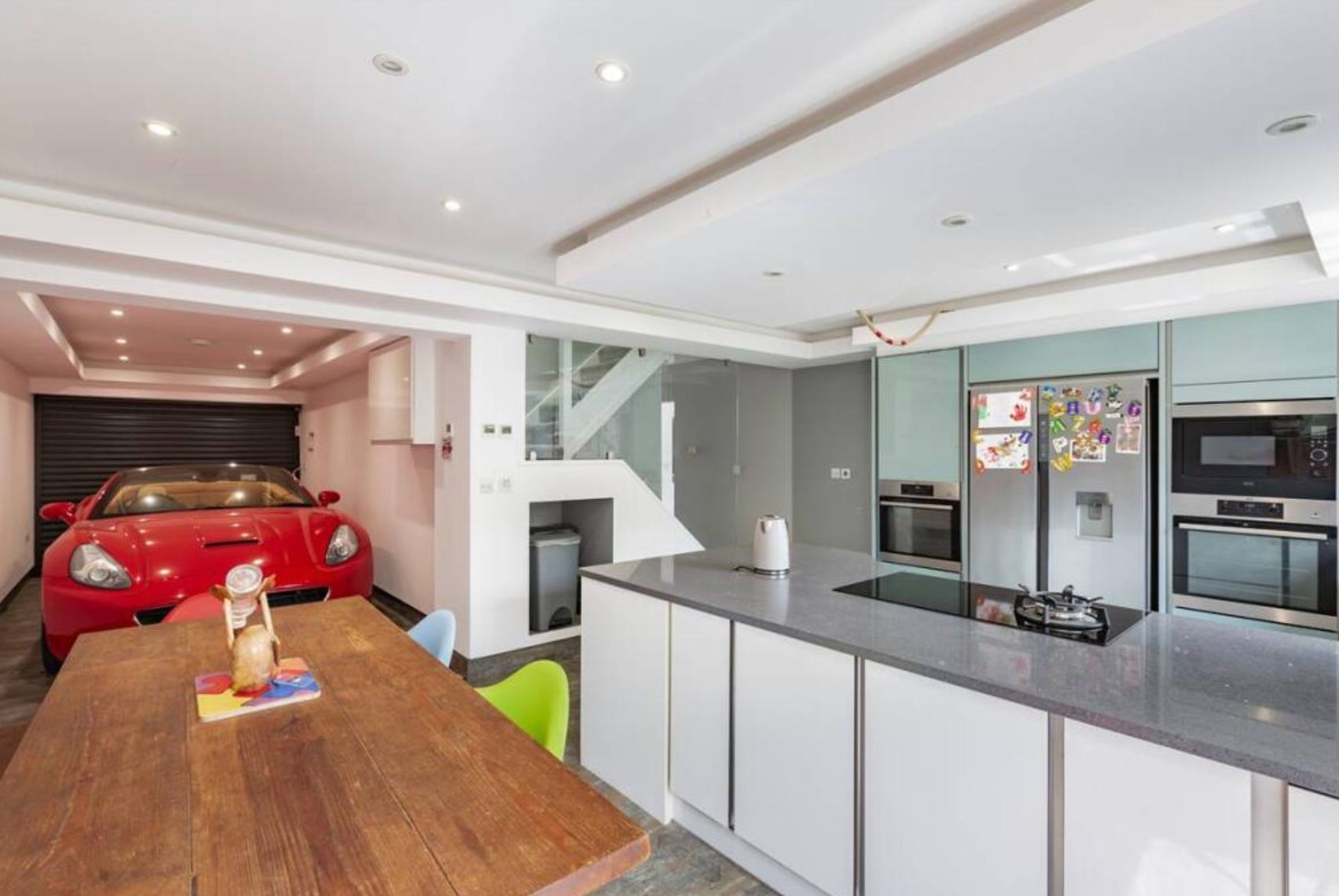 A white kitchen with a dining table - the garage is right next to it and is open to the rest of the room. There is a red car pulled in that looks like it's at the dining table.
