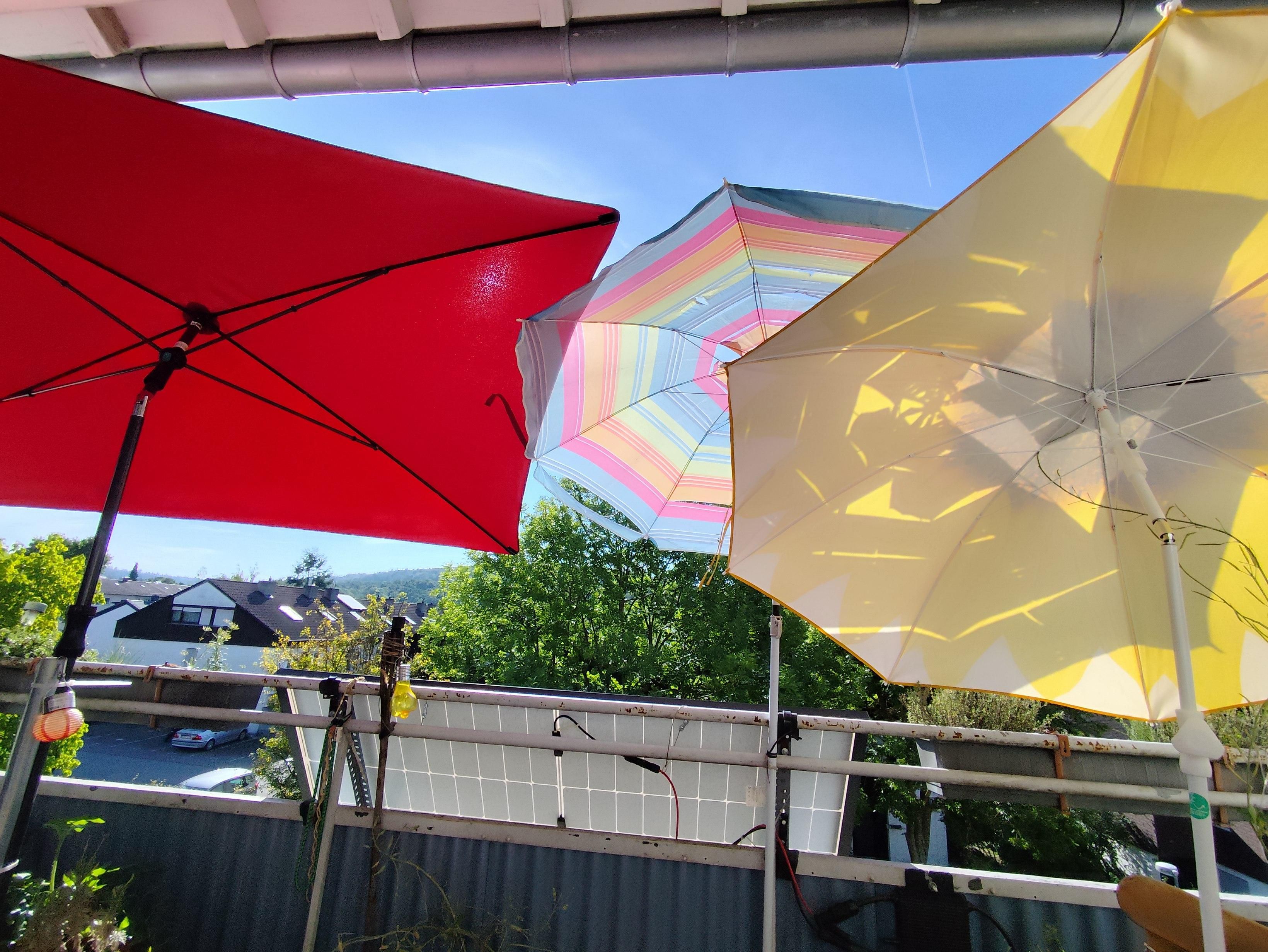Drei, zT voreinander gestaffelte bunte Sonnenschirme vor einer Balkonbrüstung, an der halb verdeckt ein Balkonkraftwerk zu sehen ist