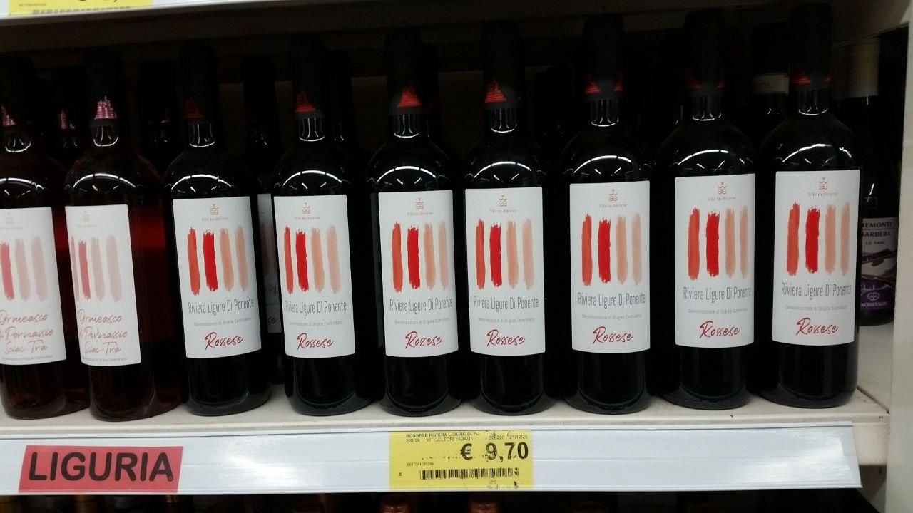 Bottles of Ligurian Rossese wine on the shelf of a supermarket.
Their labels have strips of red/pink arranged in a gradient.
The price is 9.70€/bottle.